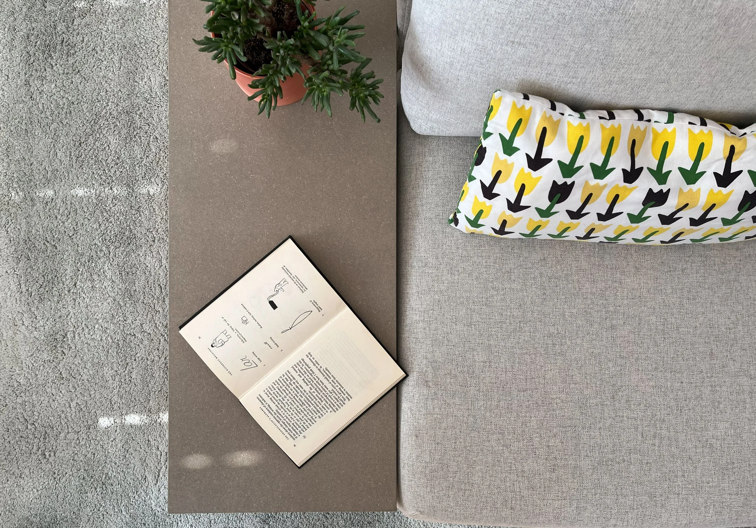 A table with a potted succulent plant, an open book, and a folded cloth with a yellow and black tulip pattern. The table is next to a gray couch with a light-colored pillow. The flooring is a speckled gray material.