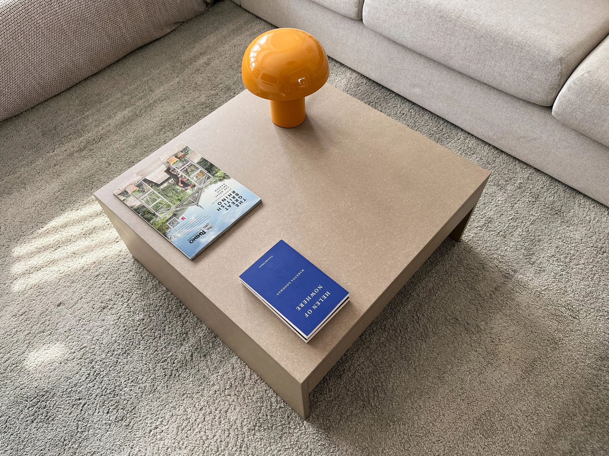 A beige rectangular coffee table with a magazine and pamphlet on top, a yellow mushroom-shaped lamp, and a grey plush sofa with textured fabric, all situated on a light grey carpet.