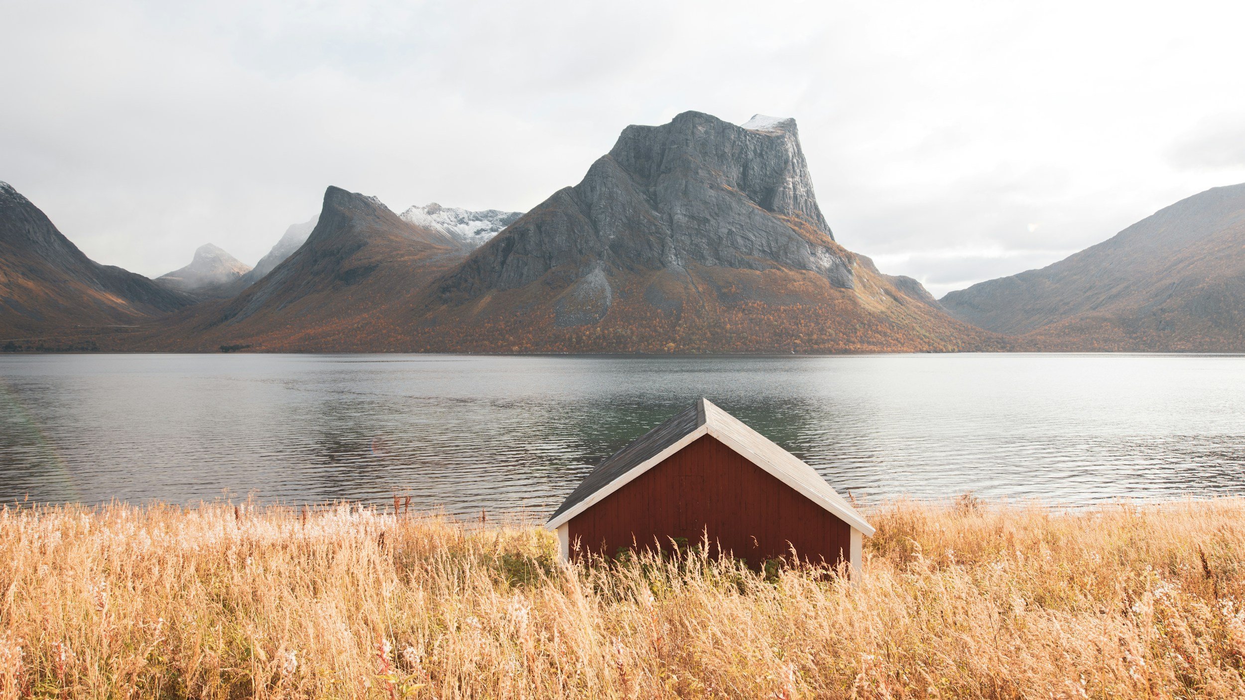 A small red house with a white roof is in the foreground, surrounded by tall, golden grass. Behind it, a calm body of water extends across the scene. Towering, rugged mountains with patches of snow are visible in the background under a cloudy sky.