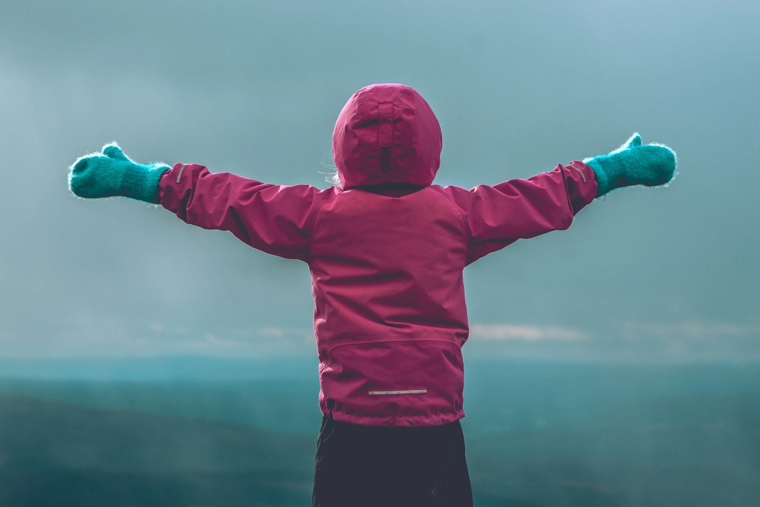 A person wearing a pink jacket, teal gloves, and a pink hood with arms outstretched in front of a cloudy sky.