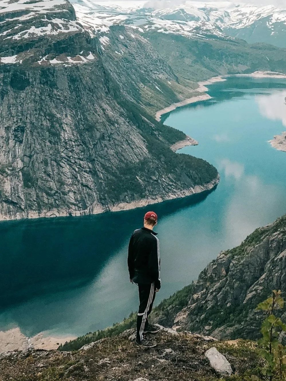 A person standing on a rocky cliff overlooking a deep valley with a large lake surrounded by steep mountains with patches of snow.