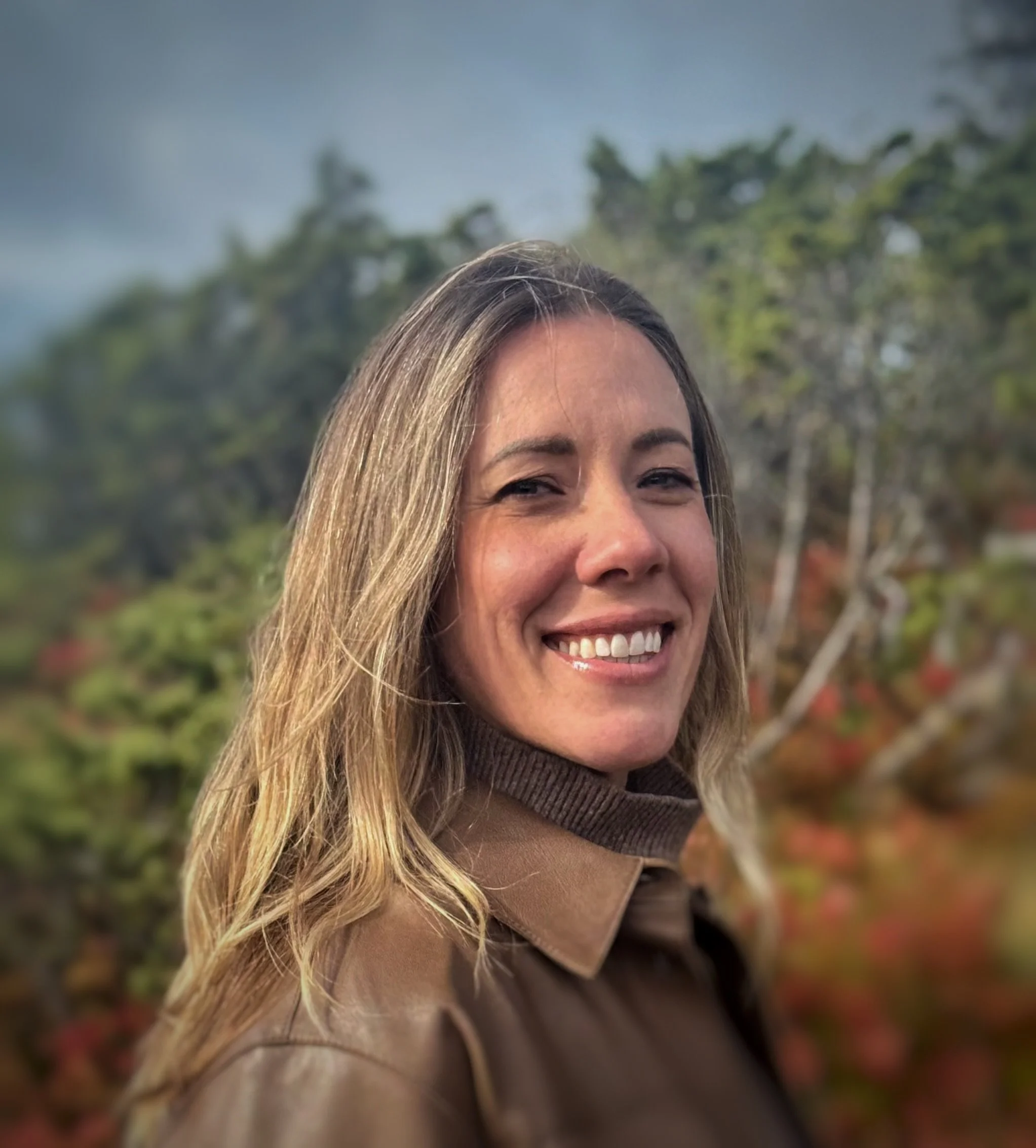A woman with long, blonde hair smiling outdoors, with trees and a cloudy sky in the background.