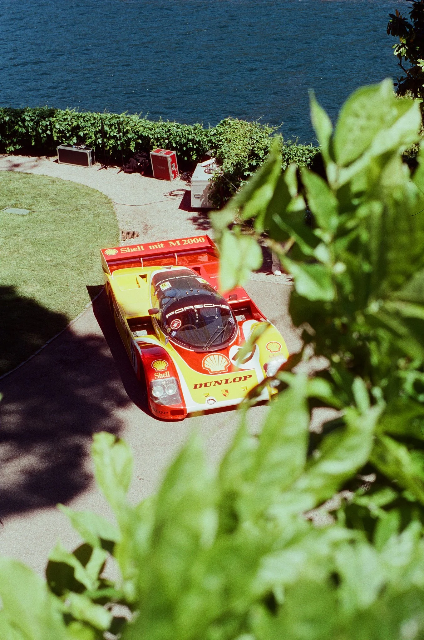 A race car with Shell and Dunlop branding parked on a driveway next to a body of water, with green leaves in the foreground.