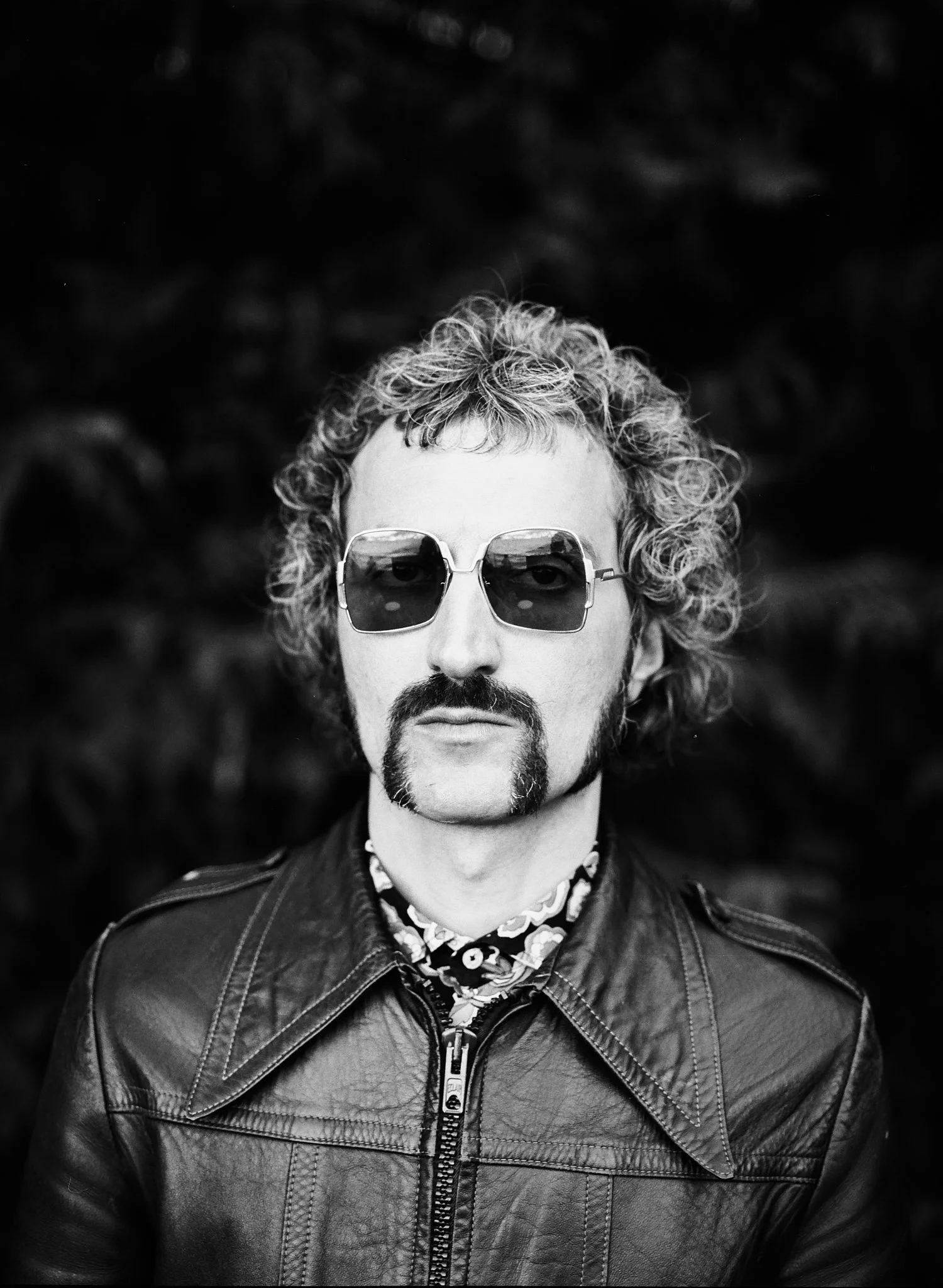 Black and white photo of a man with curly hair, wearing sunglasses, a patterned shirt, and a leather jacket, standing outdoors against a dark, blurred background.