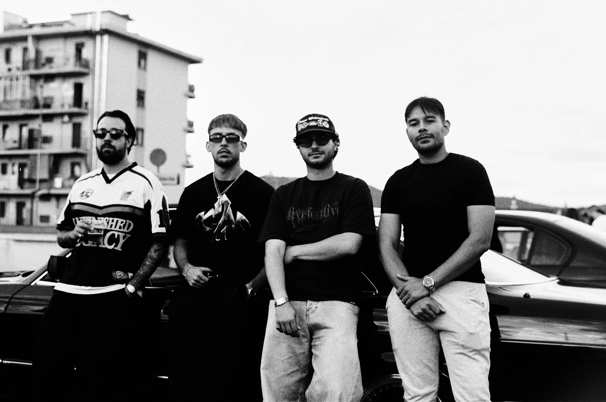 Four young men standing next to a car outdoors with a building in the background. They are wearing casual clothing and sunglasses, and are posing for the photo.