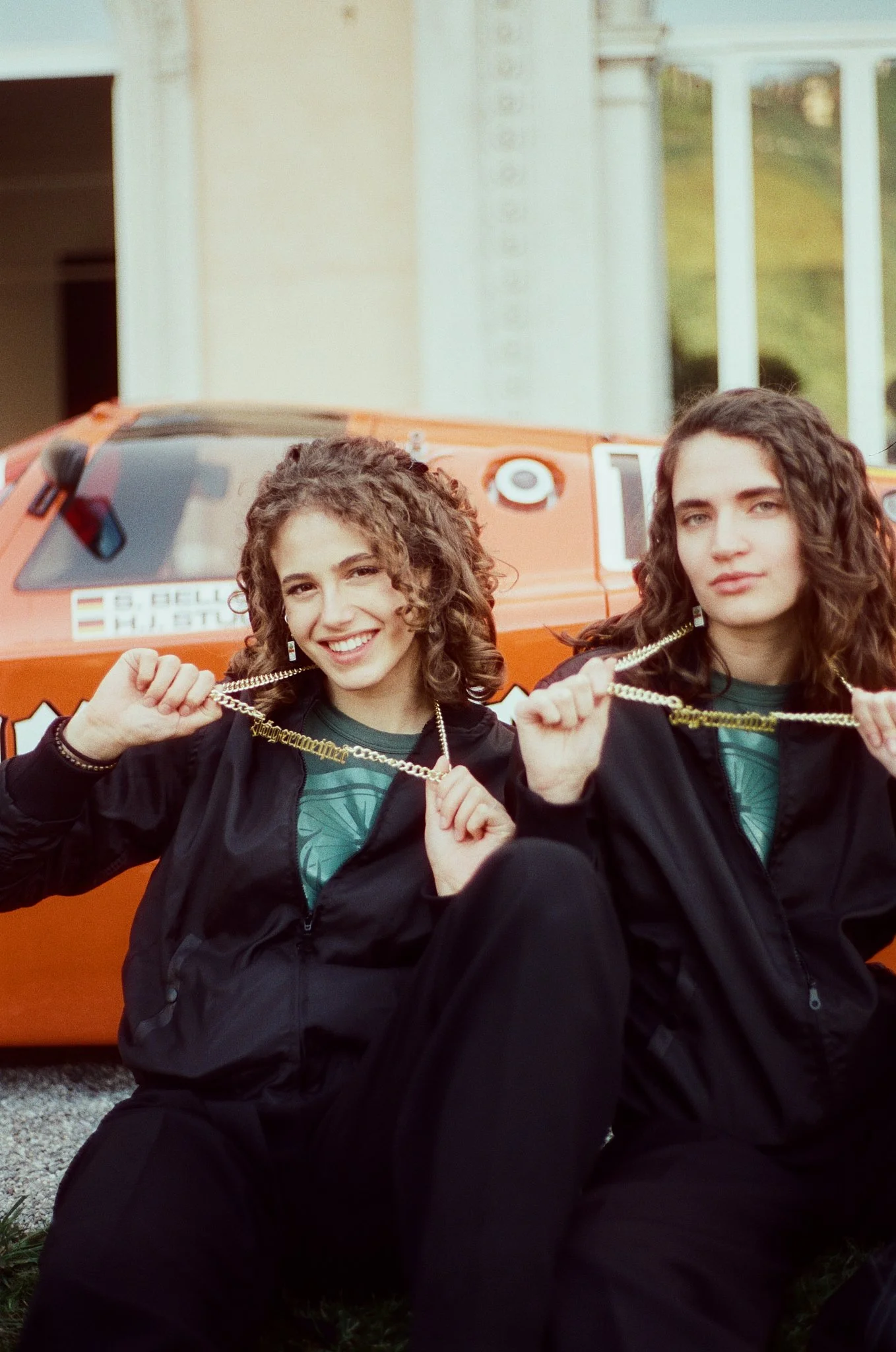 Two women with curly hair and light skin, wearing black jackets and green shirts, are sitting in front of an orange vehicle. They are holding gold chains and smiling at the camera. The vehicle has a sticker with the flag of Hungary and a license plat