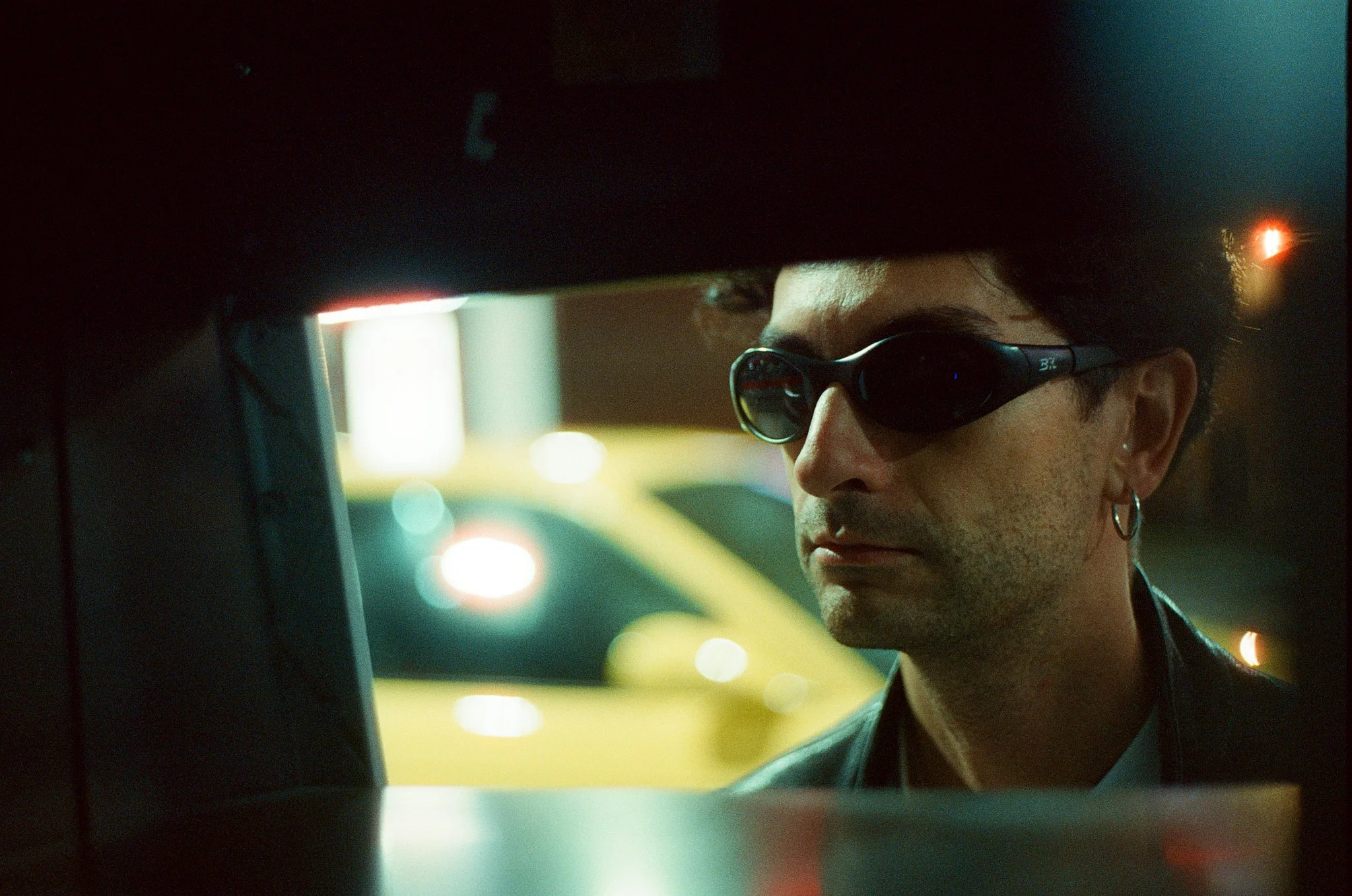 Close-up of a person wearing sunglasses, sitting in a car at night, with blurred city lights in the background.