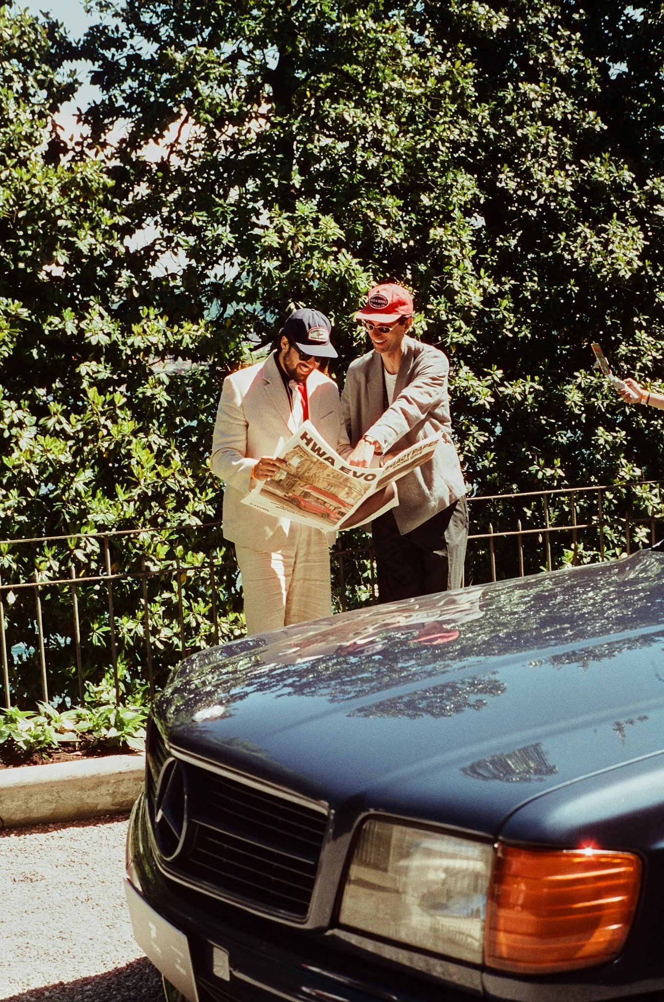 Two men, dressed in suits and wearing caps, are standing outdoors behind a black Mercedes-Benz vehicle with a reflection of trees on its hood. They are looking at a newspaper together, smiling, in front of a green leafy background.