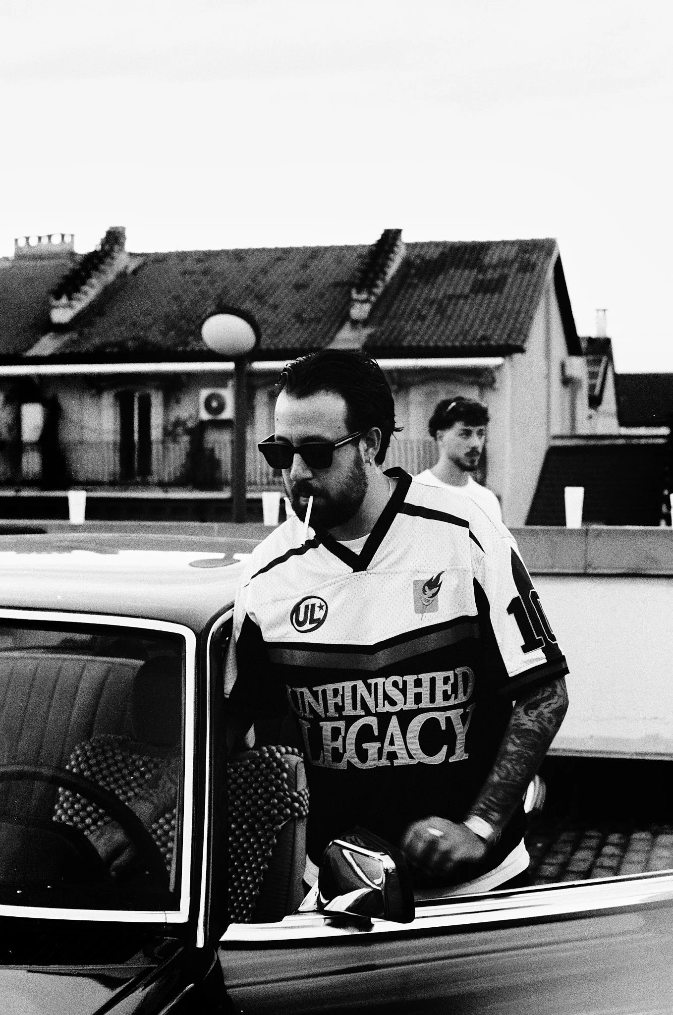 A man with dark hair, beard, and sunglasses wearing a sports jersey with the words 'UNFINISHED LEGACY' standing next to a vintage car. Another man with curly hair and mustache in the background.