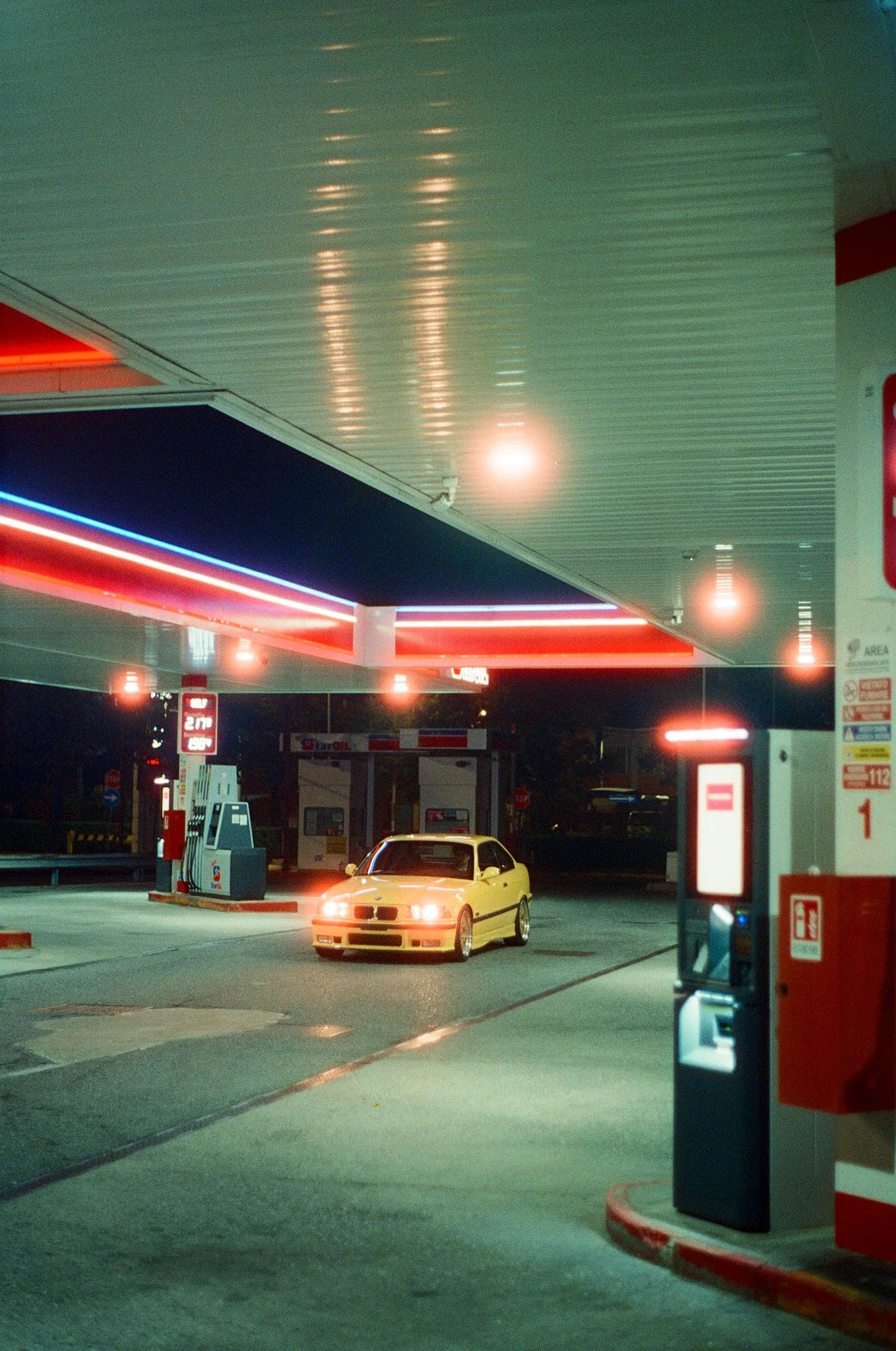 Yellow sports car parked at a gas station under fluorescent and neon lights at night.