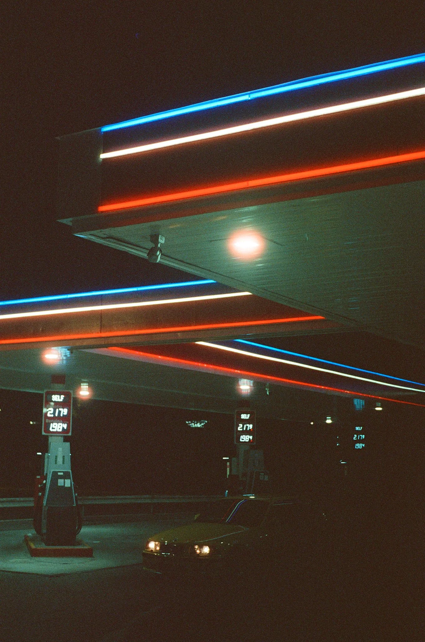 Nighttime view of a gas station with neon lights in red, white, and blue. Gas pumps are visible with digital price displays showing regular, mid-grade, and premium fuel prices.