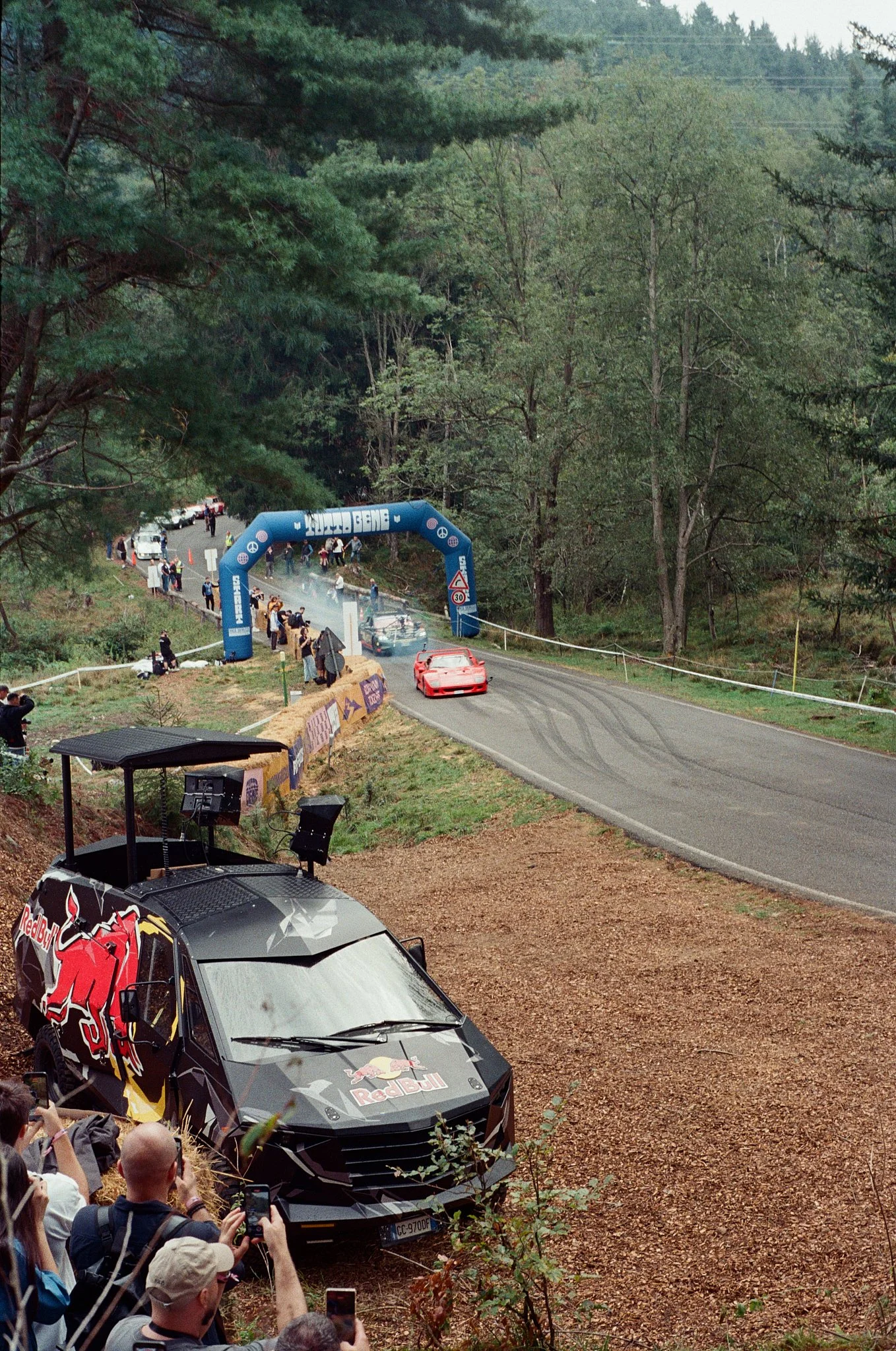 A race car with Red Bull branding on a winding road, surrounded by trees, with spectators taking photos and a starting or finishing arch in the background.