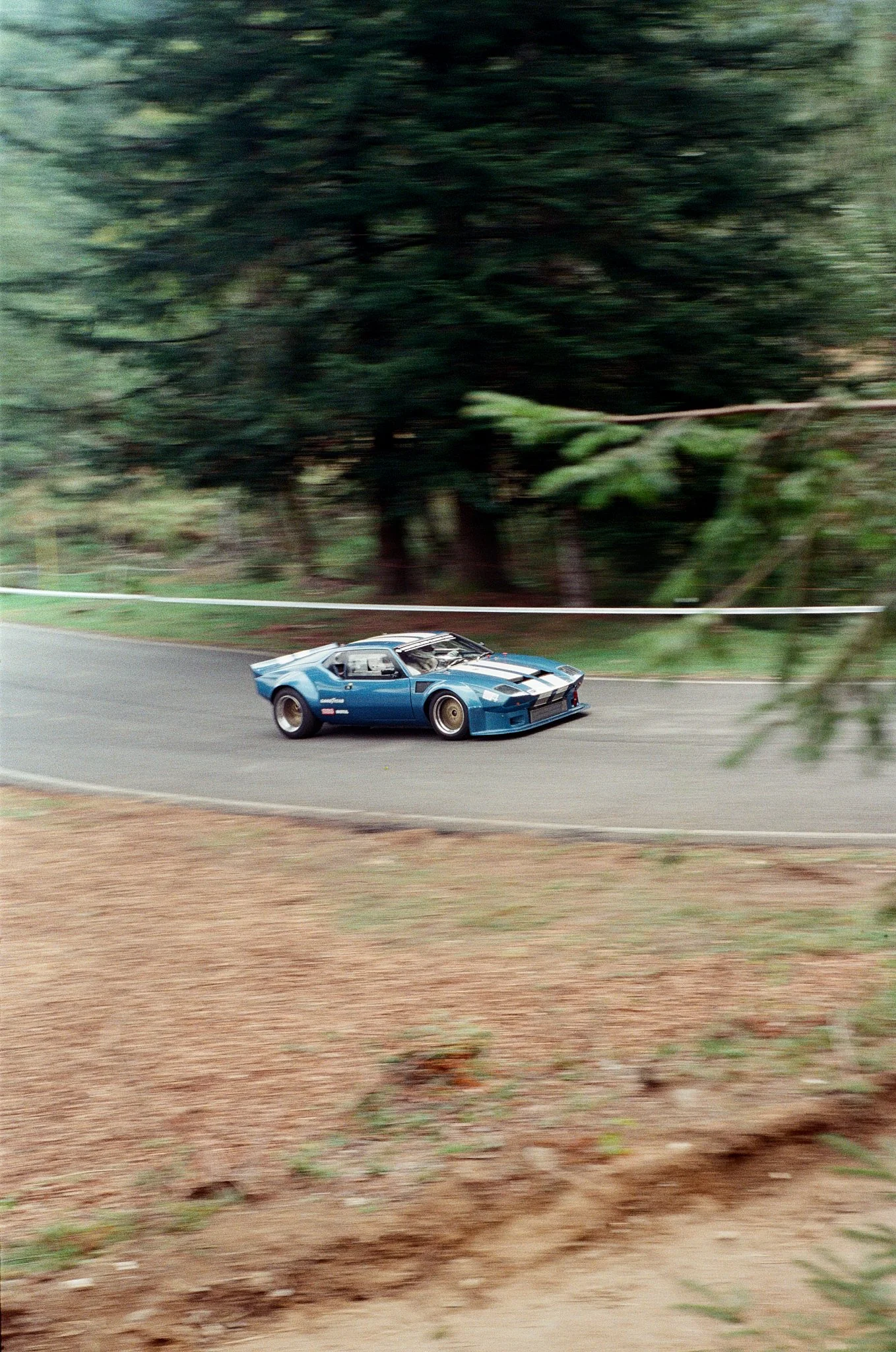 A vintage blue sports car driving on a winding road with trees and greenery in the background.