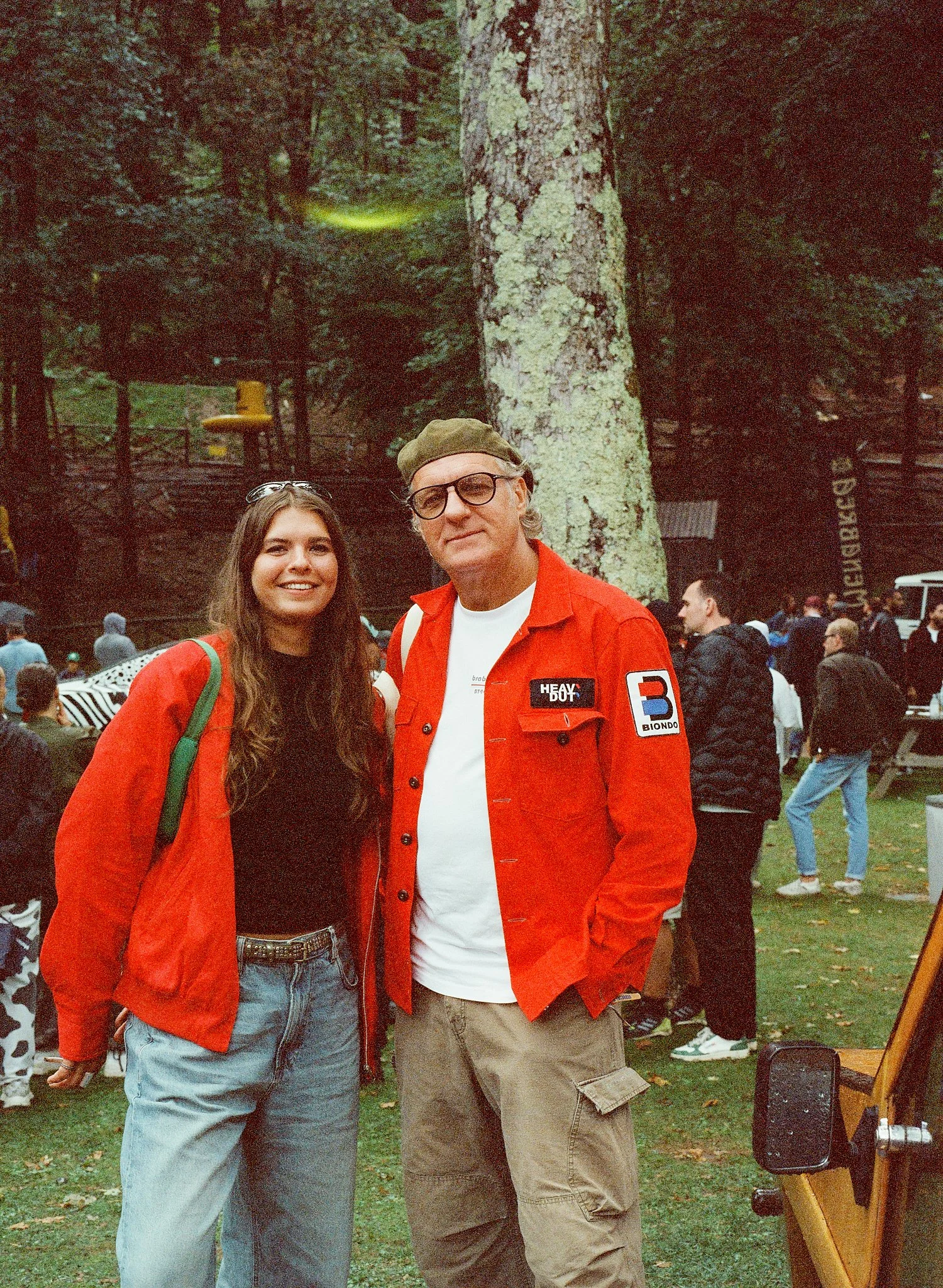 A man and a woman standing outdoors in front of a large tree, surrounded by other people at an outdoor event. The man is wearing sunglasses, a green cap, a white t-shirt, and a red jacket with patches, and the woman has long brown hair, sunglasses on