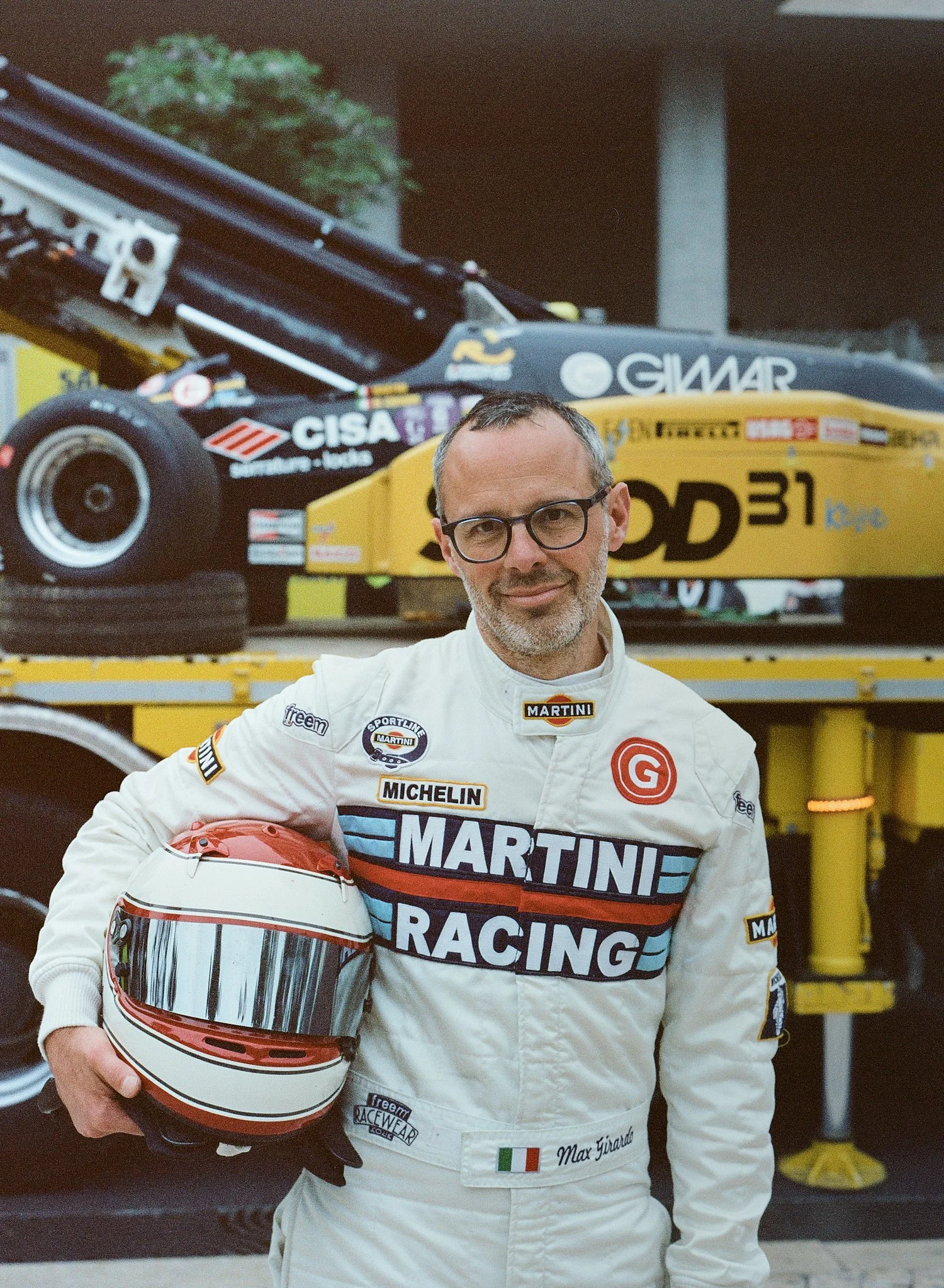 A man in a racing suit holding a white racing helmet with a red stripe, standing in front of a race car transporter. The racing suit has various sponsor logos, including Martini and Michelin, and a name tag that reads Max Girardo. The race car transp