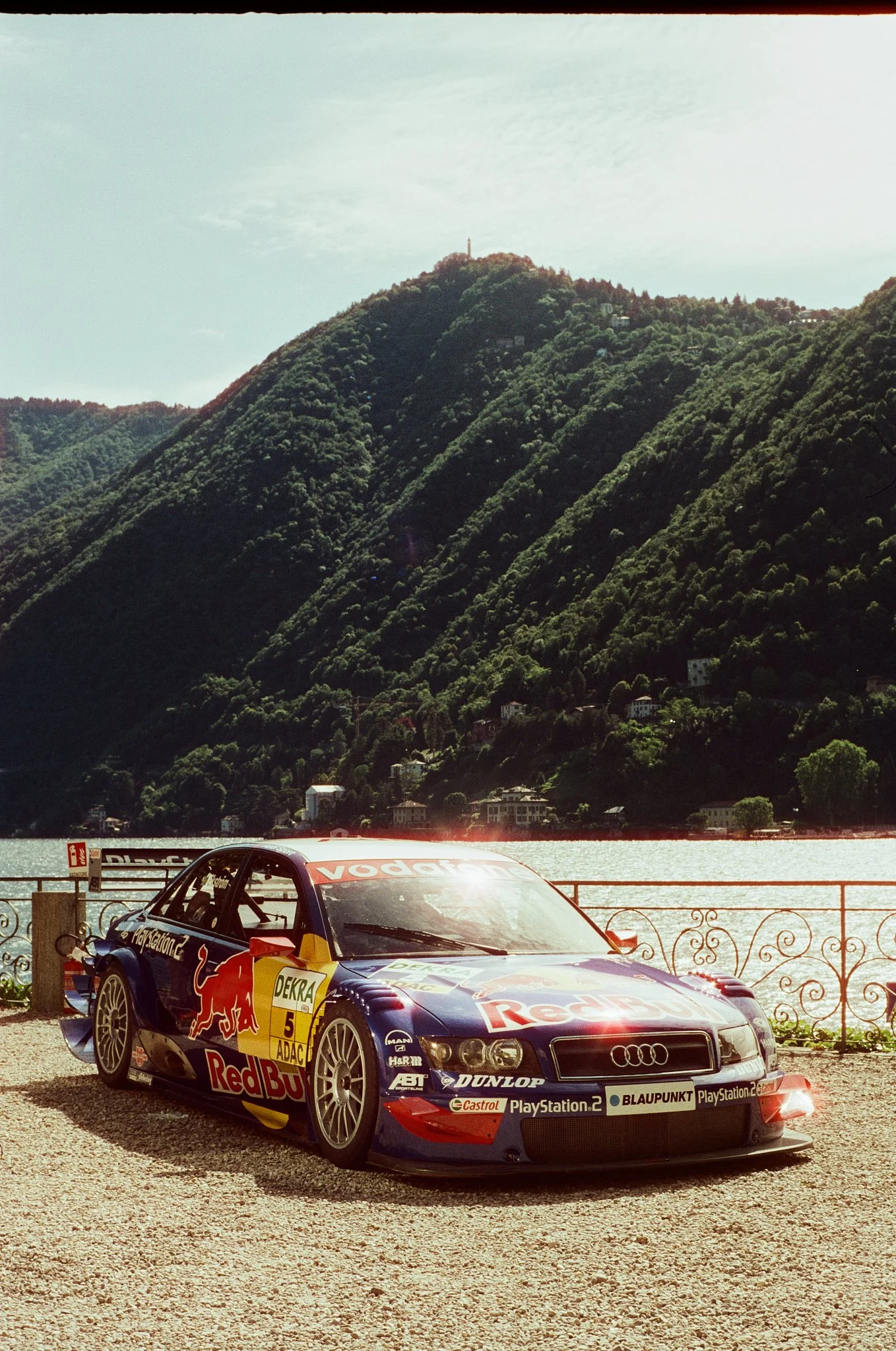 A rally car with Red Bull and Audi logos parked on a gravel surface in front of a scenic backdrop of green mountains and a lake, with houses scattered on the hillside.