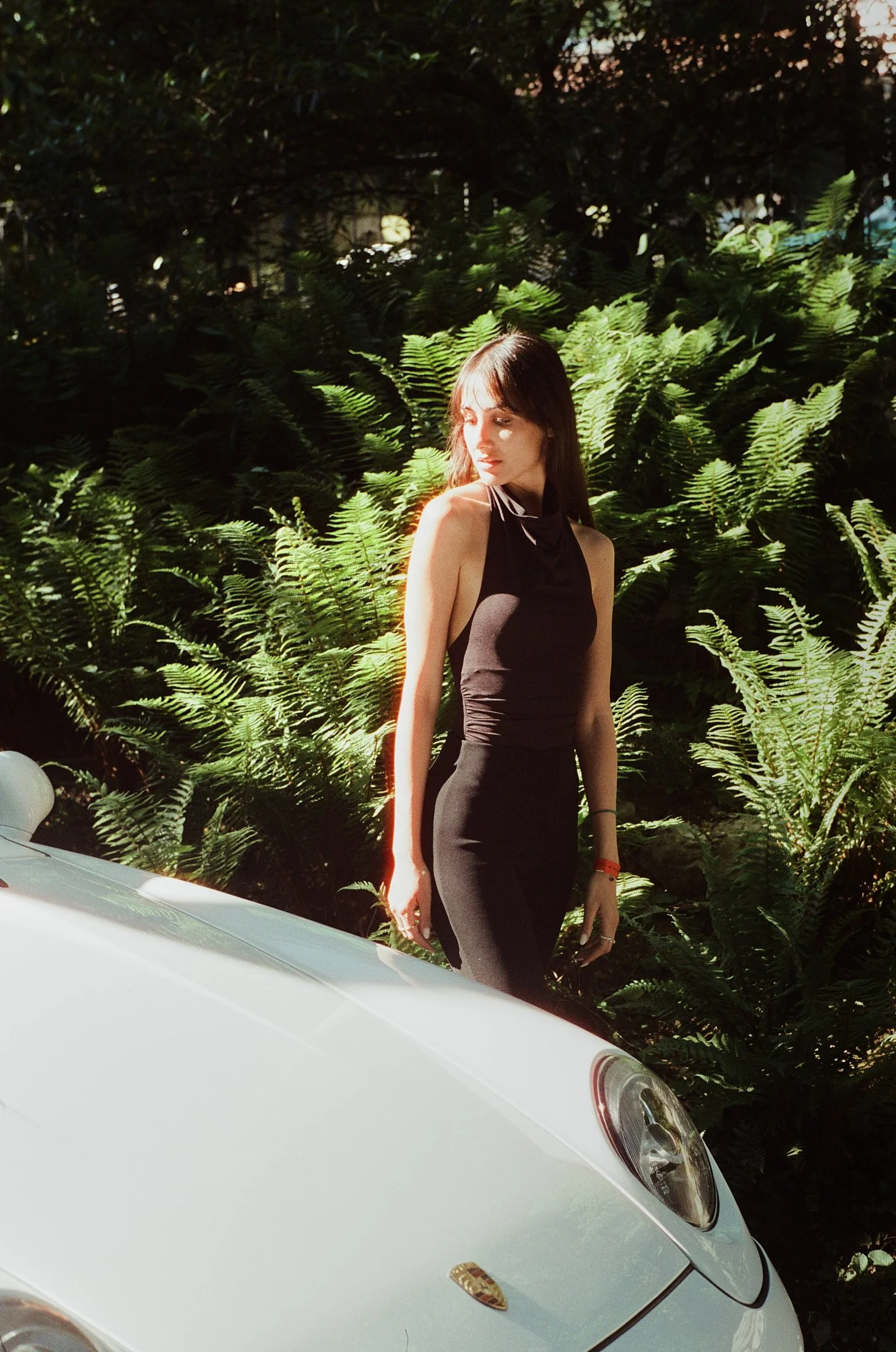 A woman with long dark hair, wearing a form-fitting black dress, is standing next to a white Porsche car. She is outdoors, with lush green ferns in the background, and sunlight highlighting her face and shoulder.