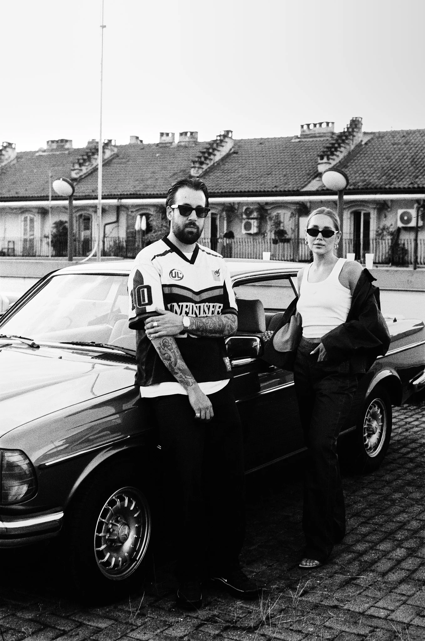 Man and woman stand next to a classic car outdoors, both wearing sunglasses. The man has a beard and tattoos on his arms, wearing a sports jersey. The woman is wearing a tank top, jacket, and sandals, with her hair tied back. Background of residentia