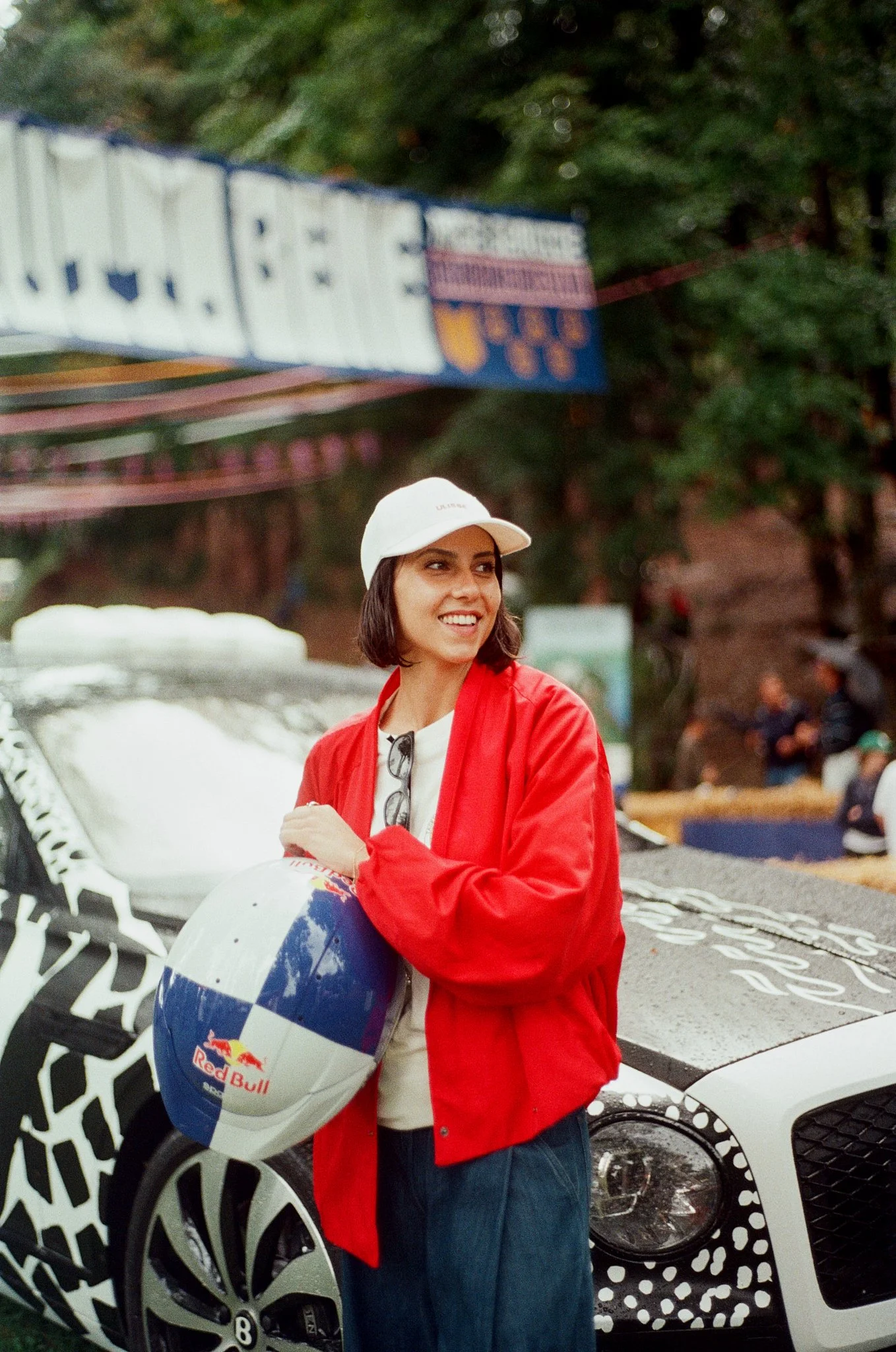 A woman smiling, wearing a white cap and red jacket, holding a blue and white Red Bull helmet, standing in front of a white sports car with a black patterned circular design on the wheels, in an outdoor setting with trees and a sign in the background.
