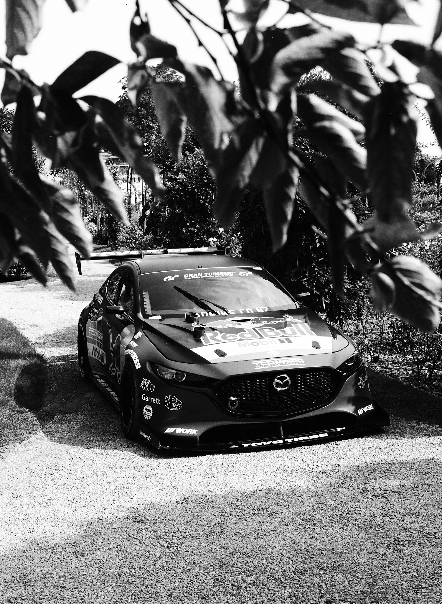 Black and white photograph of a race car with sponsor logos and a Mazda emblem, parked on a gravel surface amid trees and foliage.