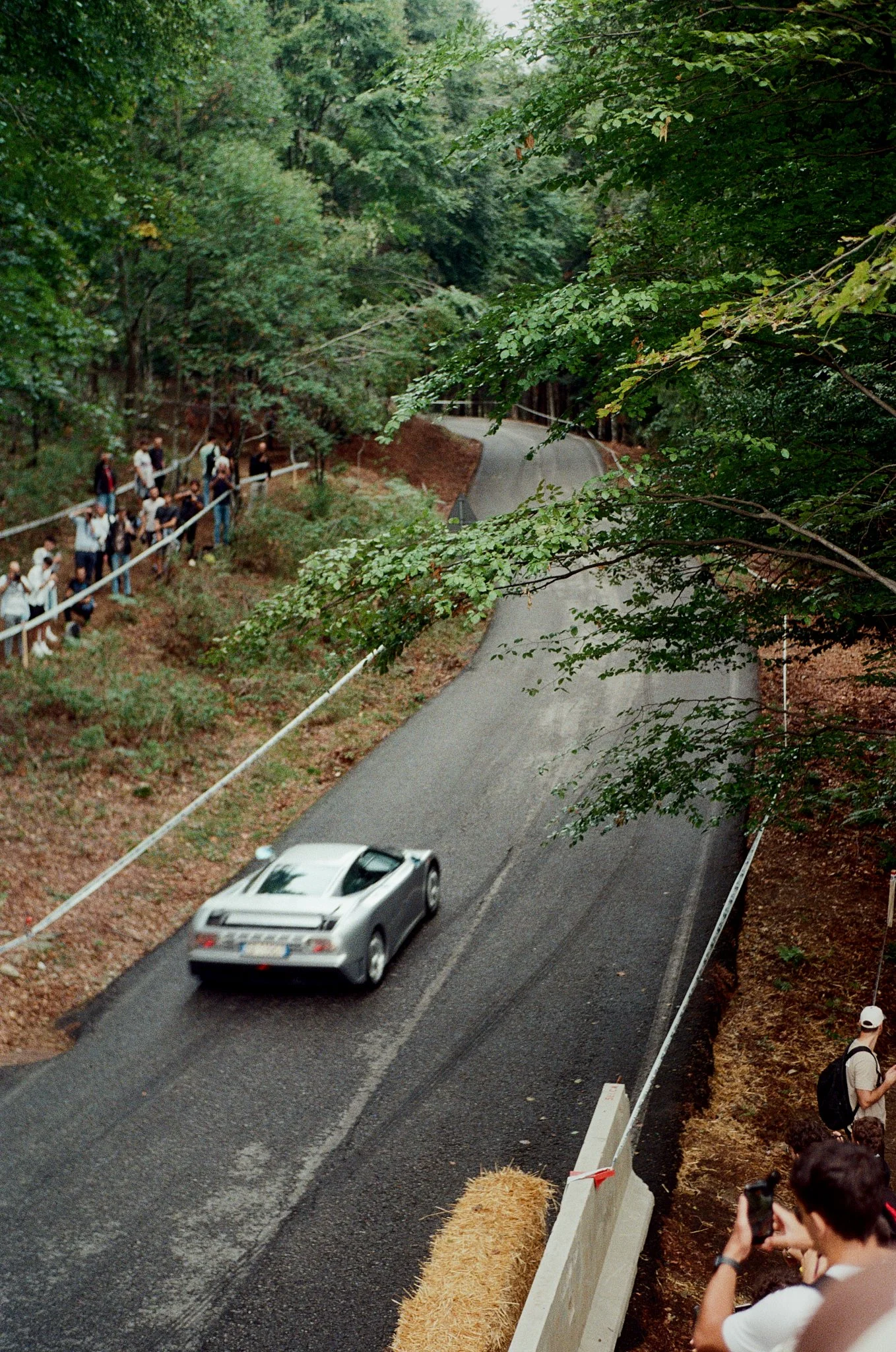 A winding road in a forested area with spectators and photographers on the sidelines, some taking pictures, a white sports car on the road, and safety barriers along the edge of the road.