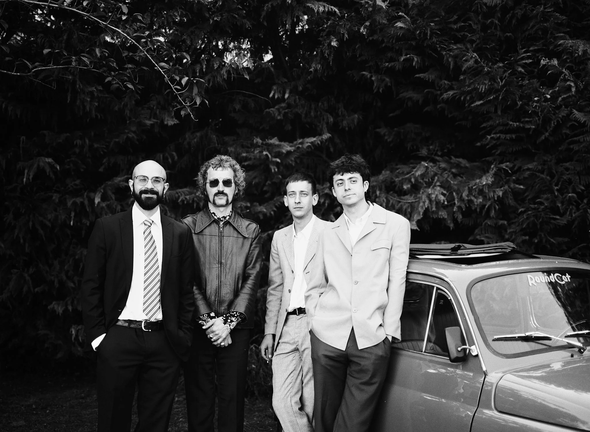 Four men standing outdoors in front of dense trees, dressed in formal and casual clothing, with part of a vintage car visible on the right.
