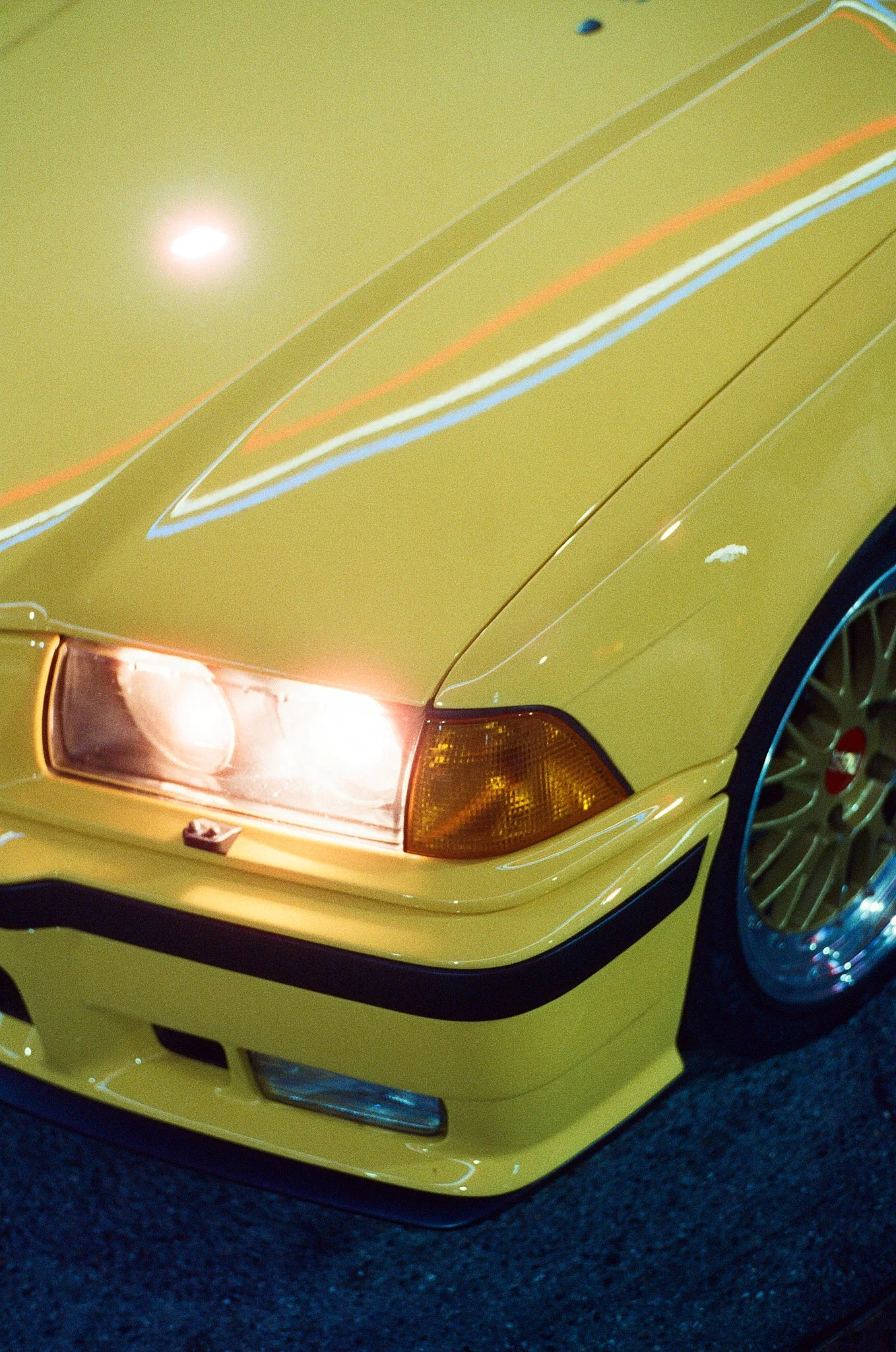 Close-up of the front of a yellow sports car, showing part of the headlight, the hood with racing stripes, and a section of the wheel.
