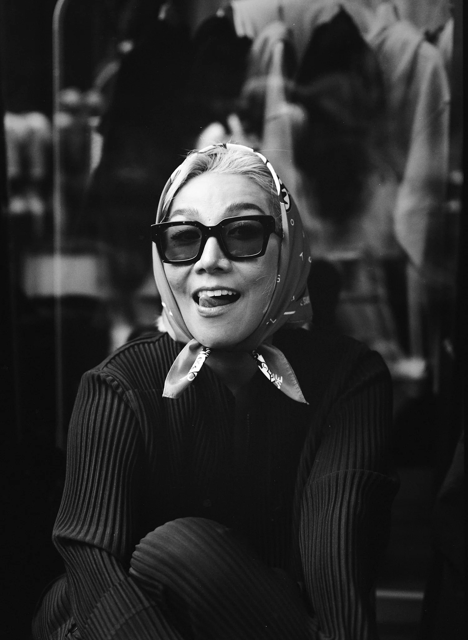 A woman with light hair, wearing large sunglasses, a headscarf, and a textured top, smiling and sticking out her tongue, viewed through a glass reflection in black and white.