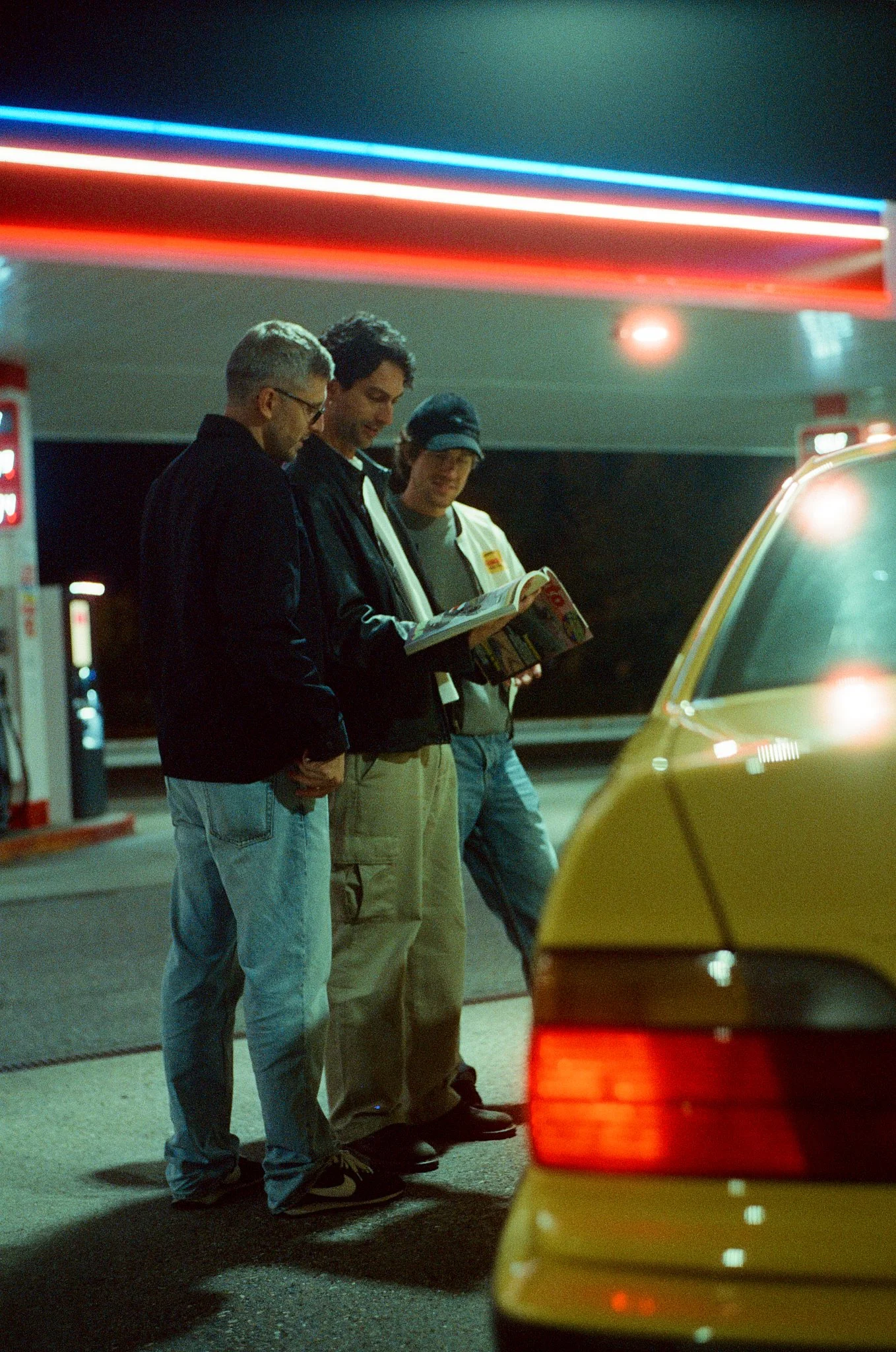 Three people standing at a gas station at night, looking at something together. A yellow car is partially visible in the foreground, and a gas pump is on the left side of the image. The scene is illuminated by neon lights.