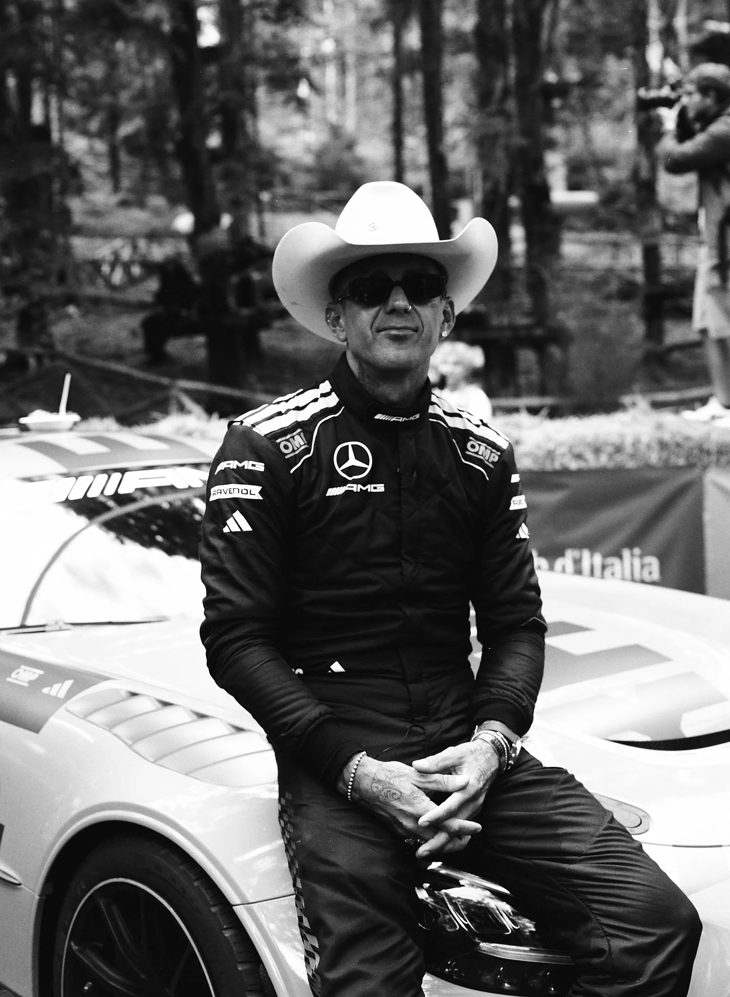 A man wearing sunglasses, a cowboy hat, and a racing suit with logos, sitting on the hood of a sports car in an outdoor setting. The background has trees and other people.
