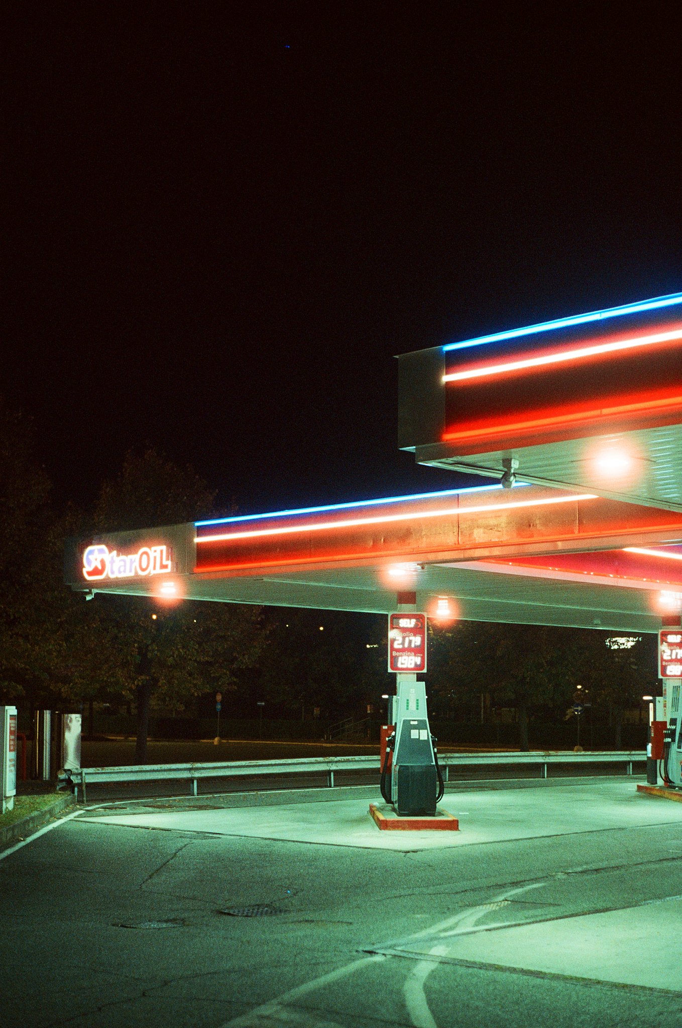 Nighttime scene at a gas station with neon lights, empty parking lot, fuel pump in the foreground, and illuminated signs with fuel prices.