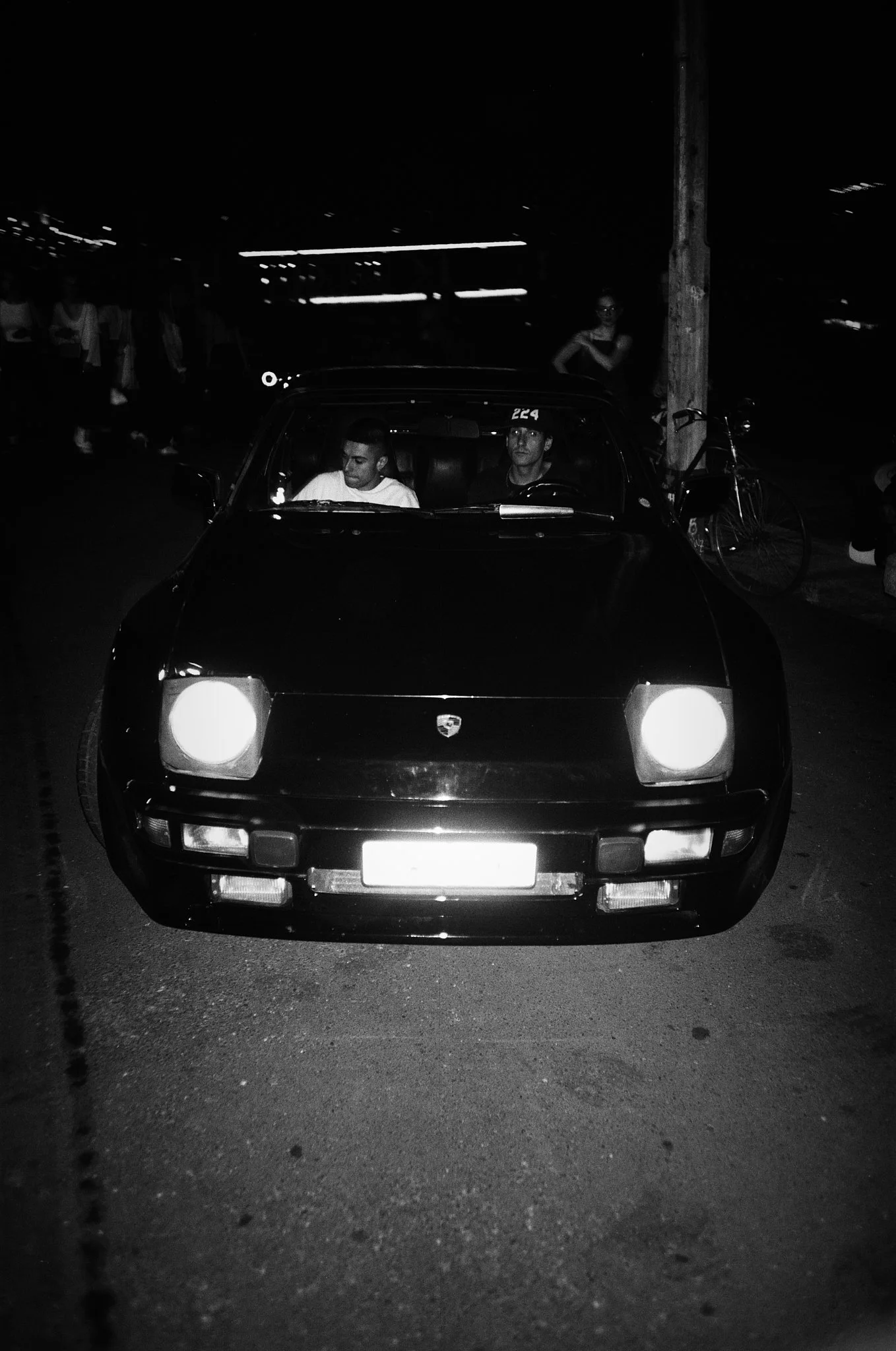 A classic black Porsche 944 sports car parked on the street at night with two men inside, one in the driver's seat and the other in the passenger seat. There are people and a bicycle nearby in a dimly lit area.