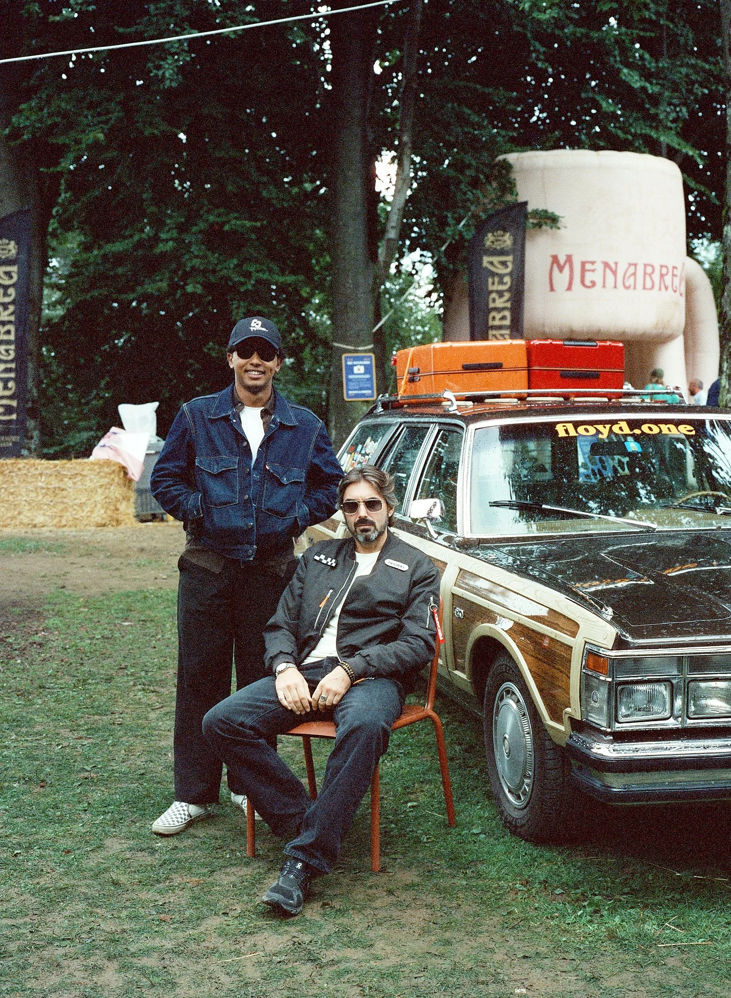 Two men pose outdoors in front of a vintage car, with one seated on a chair and the other standing, surrounded by trees and equipment.