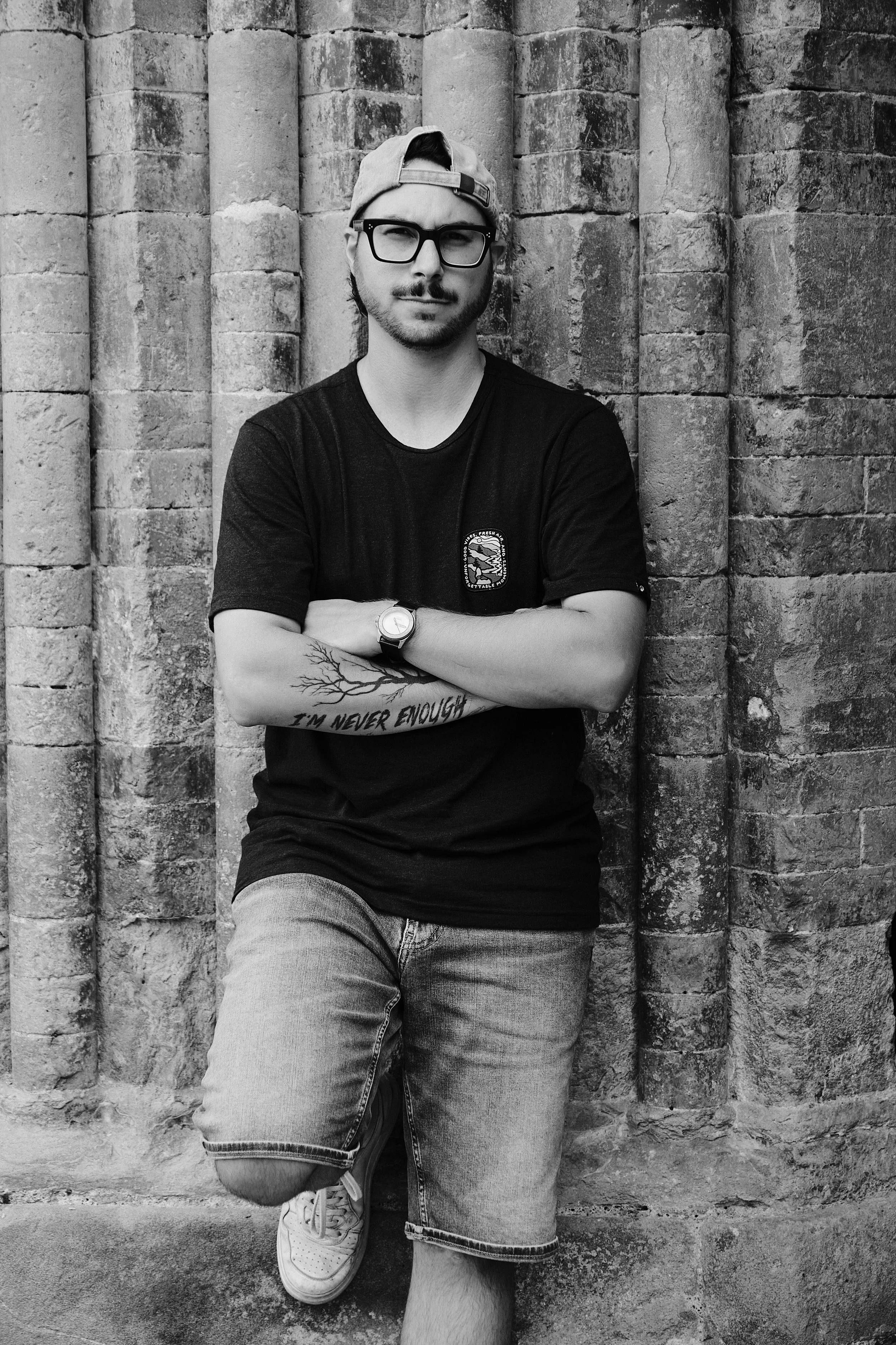 A young man with glasses, a backwards cap, and a beard, standing in front of a brick wall. He is crossing his arms, one leg slightly raised, wearing a black t-shirt and jeans, with tattoos and a watch on his left arm, and a serious expression.