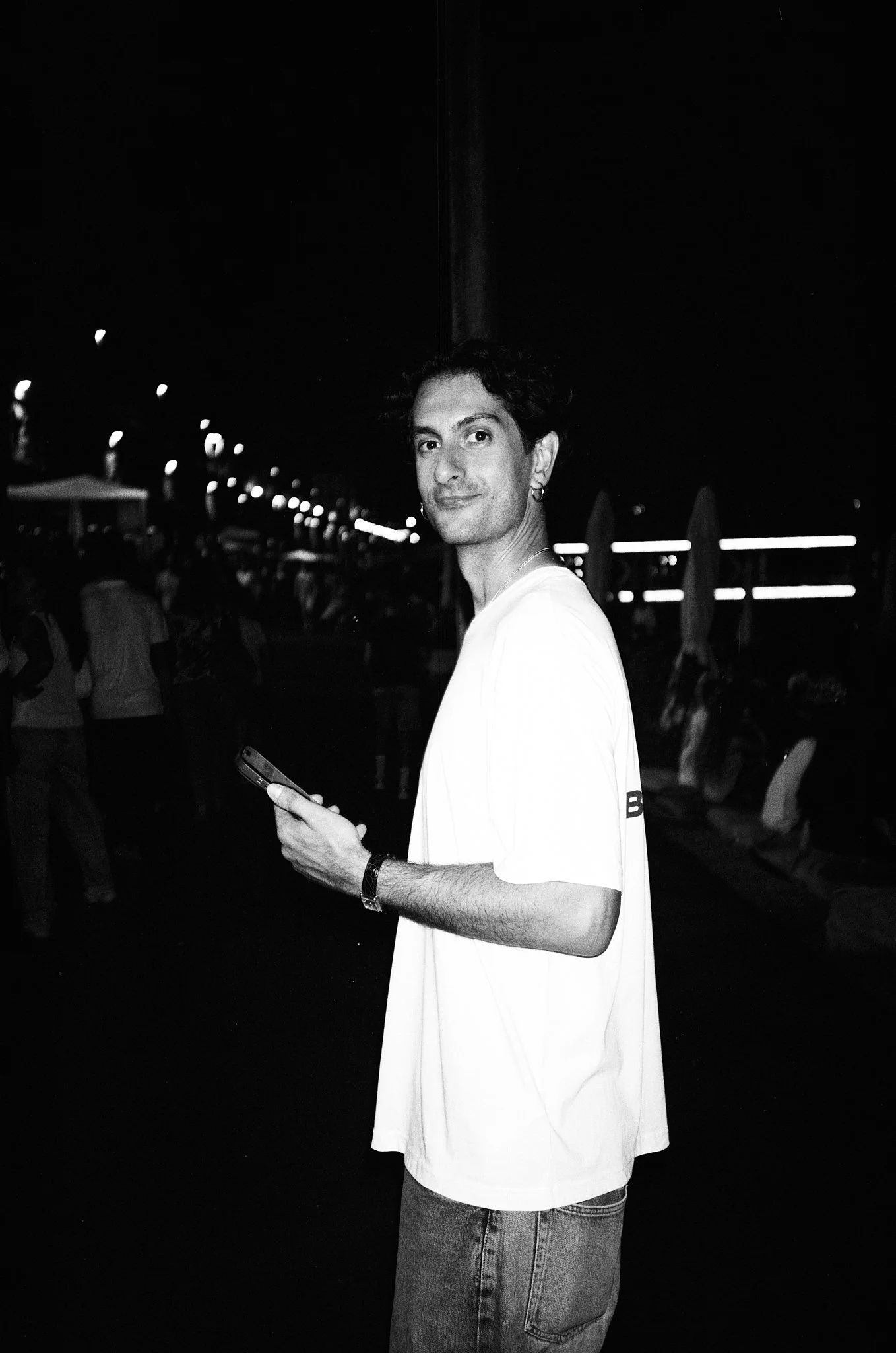 A young man with curly hair, wearing a white T-shirt and jeans, stands outdoors at night holding a smartphone. The background features blurred lights and other people.