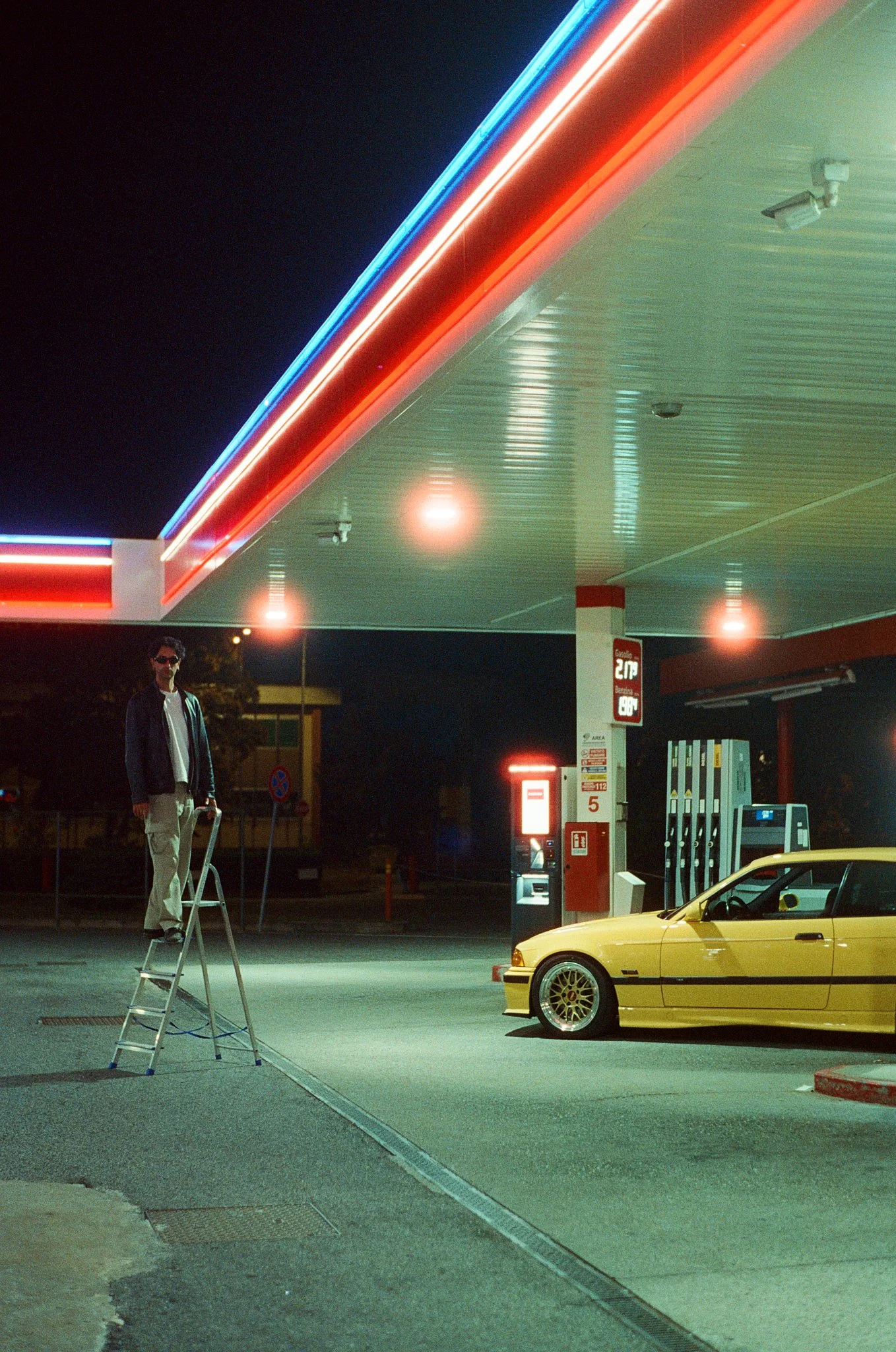 Night scene at a gas station with a yellow sports car parked at the pumps, a man standing on a ladder, and neon lights illuminating the area.