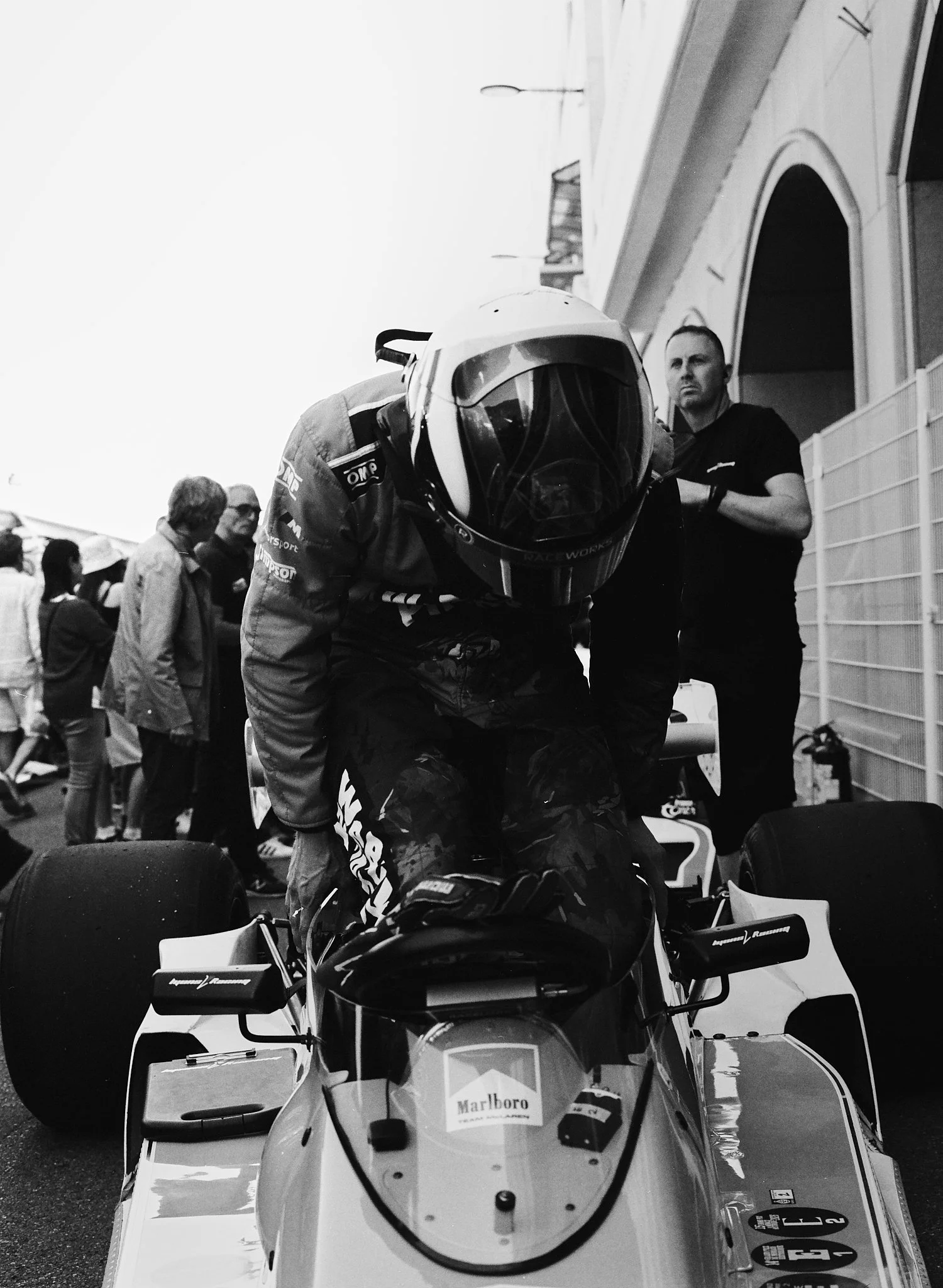 Black and white photograph of a race car driver wearing a helmet, leaning forward on a race car, with people in the background standing and observing in a paddock or garage area.