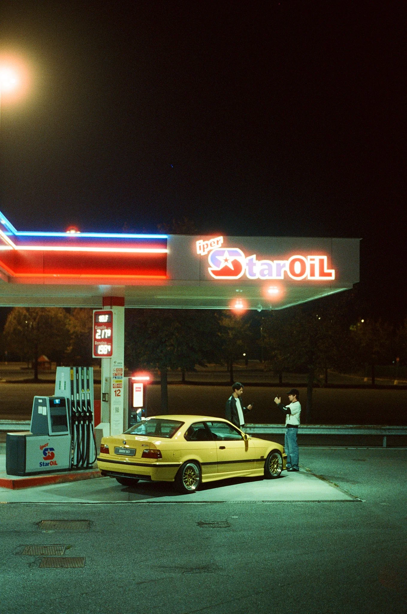 Nighttime at a gas station with a yellow car parked at a pump. Two men are standing near the car, engaged in conversation, with one showing something on his phone. The gas station's sign reads 'Star OIL,' and the canopy has neon lights. The fuel pric