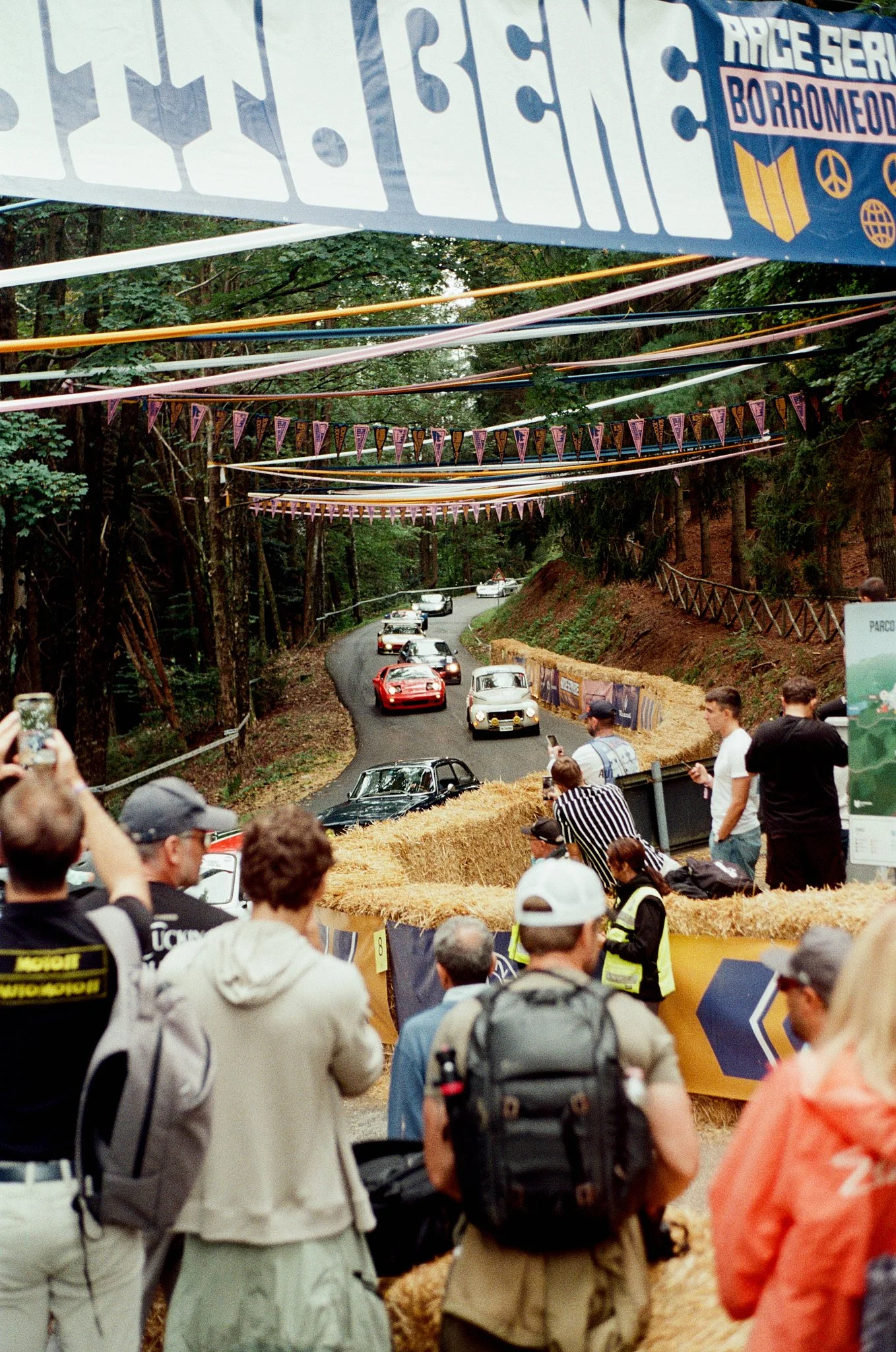 A street with classic cars driving around a bend, surrounded by trees. People are gathered along the sides, taking photos and watching. Decorations like banners and colorful flags hang overhead, indicating a car event or race.