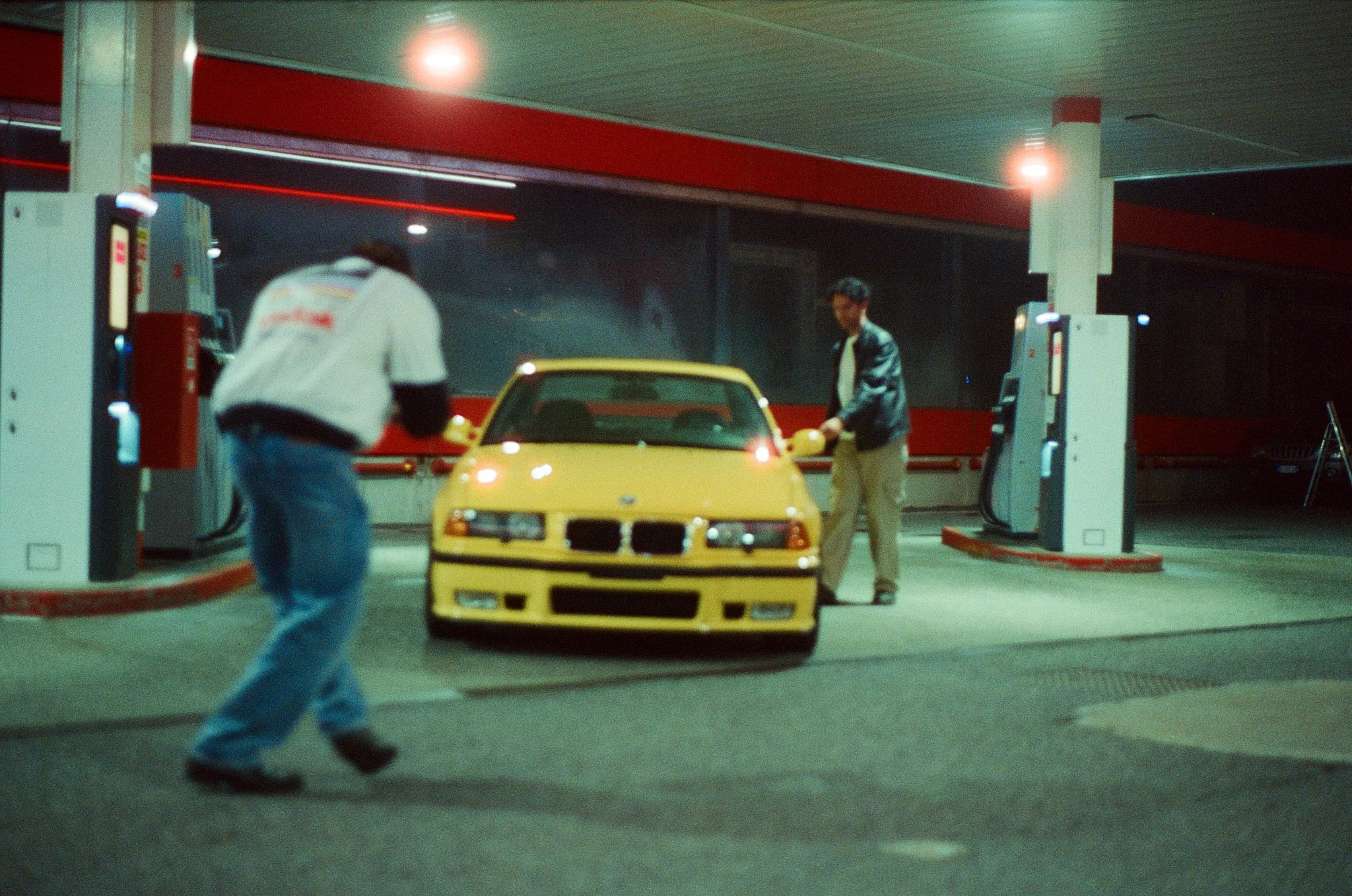 A yellow sports car at a gas station being filled with fuel by a man in a leather jacket, with another person walking past in the foreground.