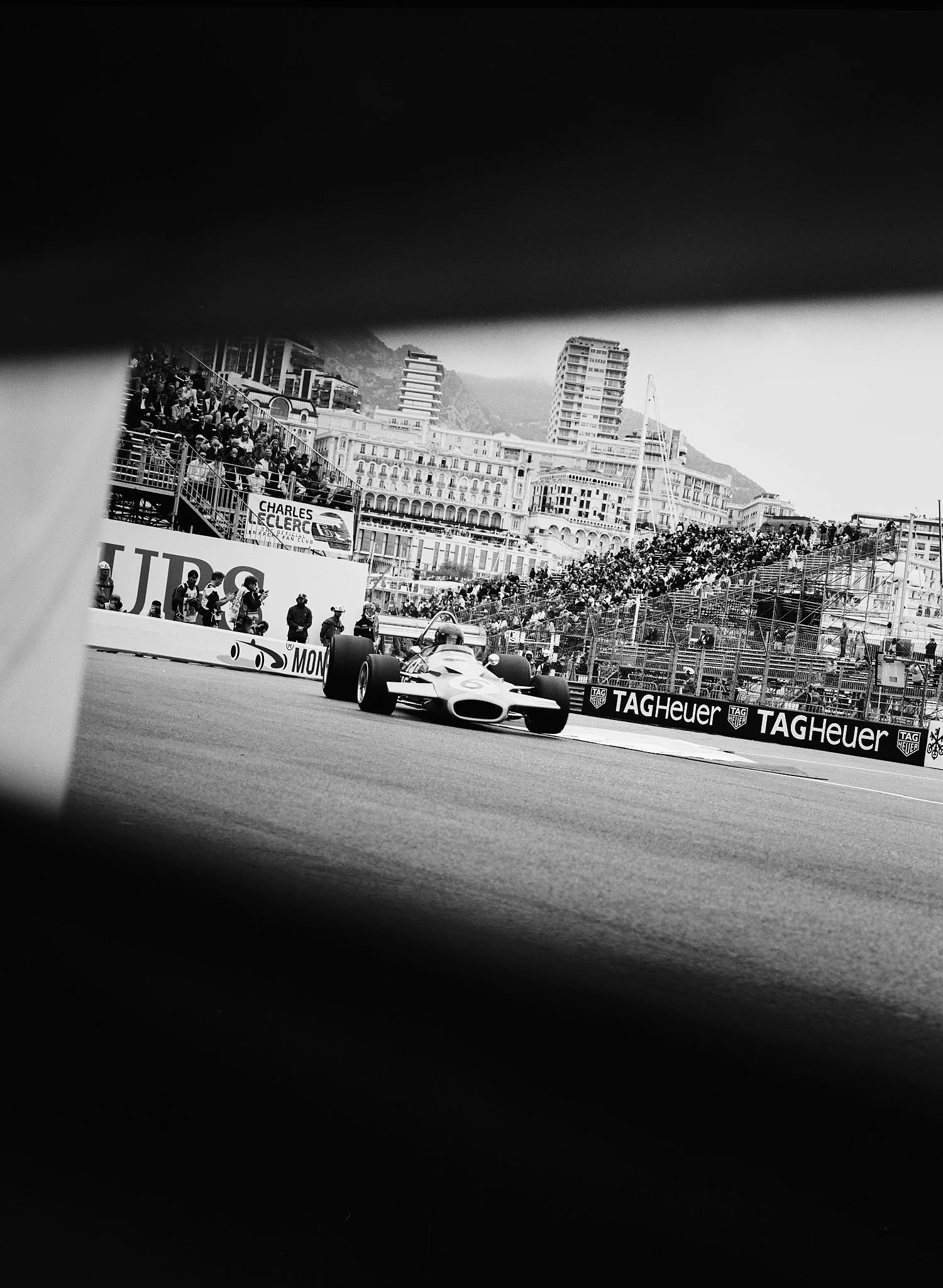 A race car speeding on a track with spectators in the stands and city buildings in the background, captured in black and white.
