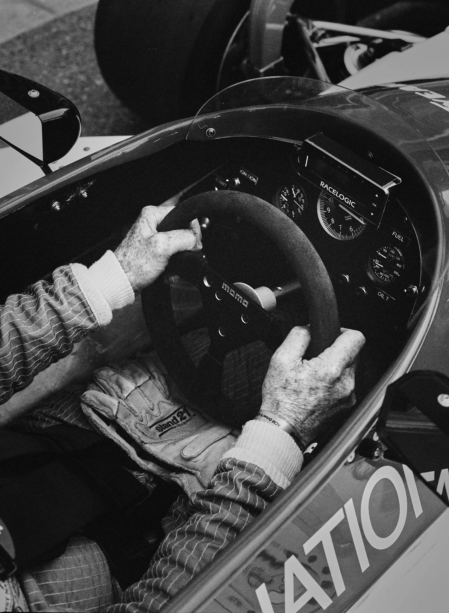 A person in racing gear sitting in a race car, holding the steering wheel. The dashboard shows gauges, and there is a digital display with the text 'RACELLOGIC'. The photograph is black and white.