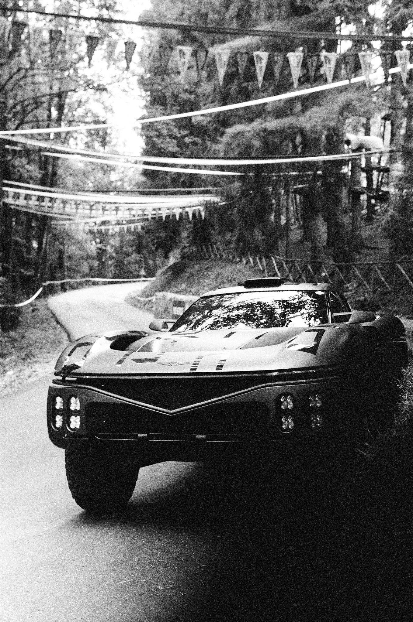 A black and white photograph of a race car parked on a road surrounded by trees. The front of the car is prominently shown with detailed headlights and a sleek design. Overhead, bunting banners are strung across the scene.