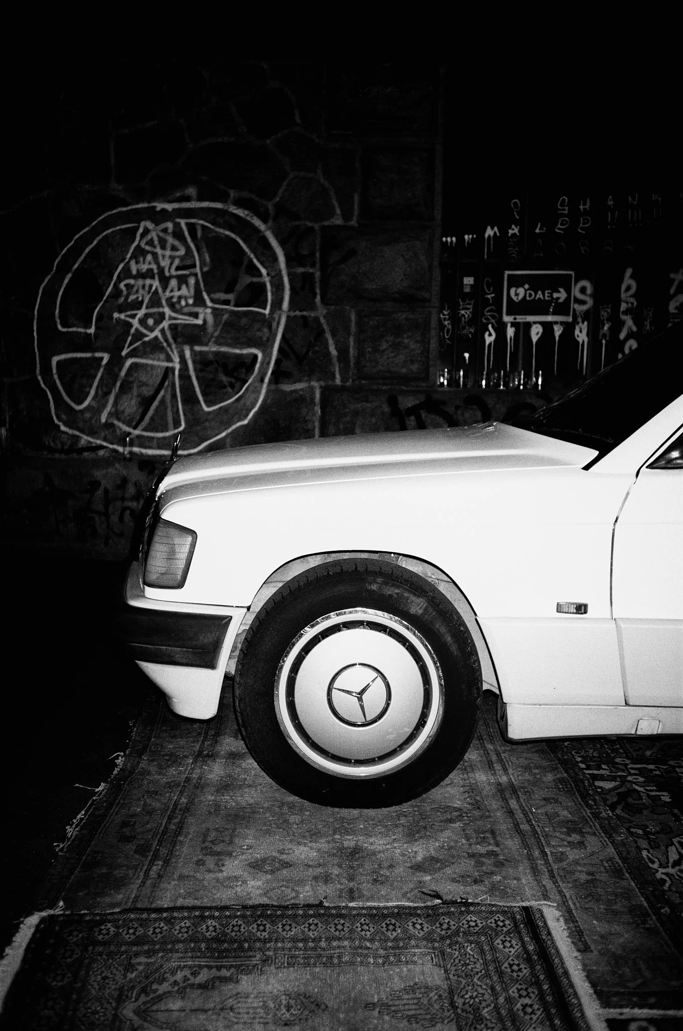 A white Mercedes-Benz car parked indoors on a Persian-style rug. The background features dark graffiti and artwork on a brick wall, including a peace sign, text, and symbols.