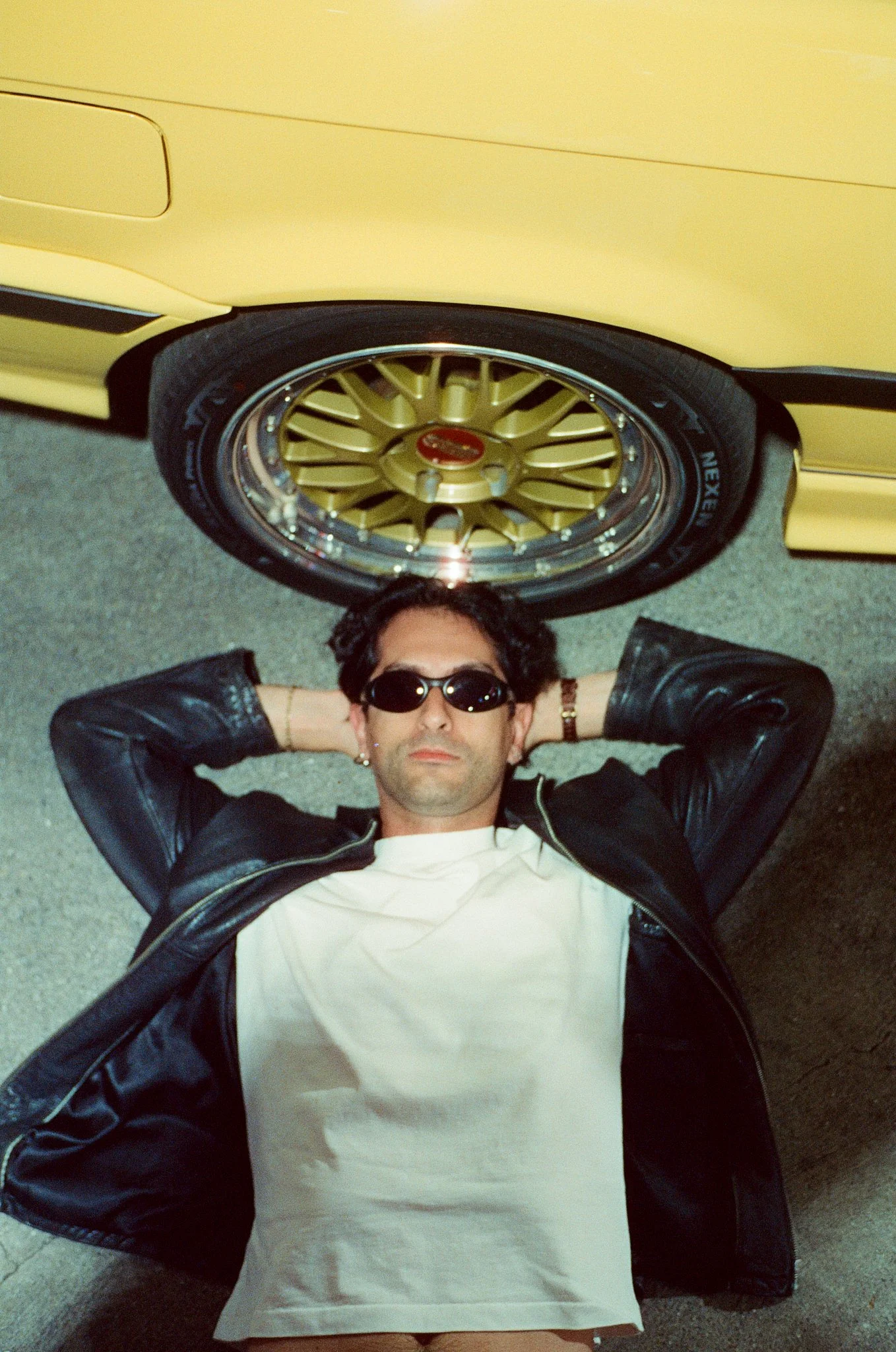 A person lying on the ground with their hands behind their head, wearing sunglasses, a white t-shirt, and a black leather jacket, with a yellow car and gold wheels in the background.