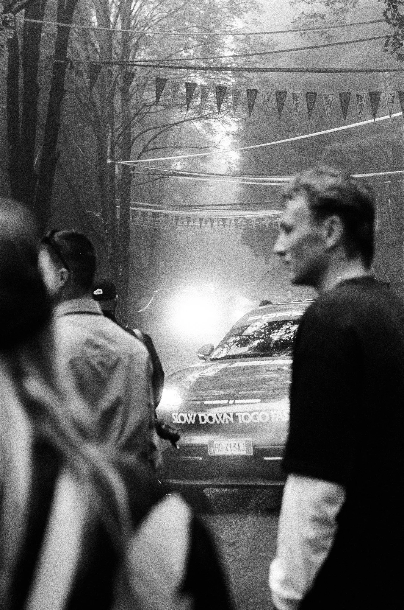 Black and white photo of a traffic scene at dawn or dusk, with two women in conversation in the foreground. Cars are visible, with one showing a partial license plate. Trees line the street with early morning or late evening sunlight filtering through, and festive banners hang overhead.