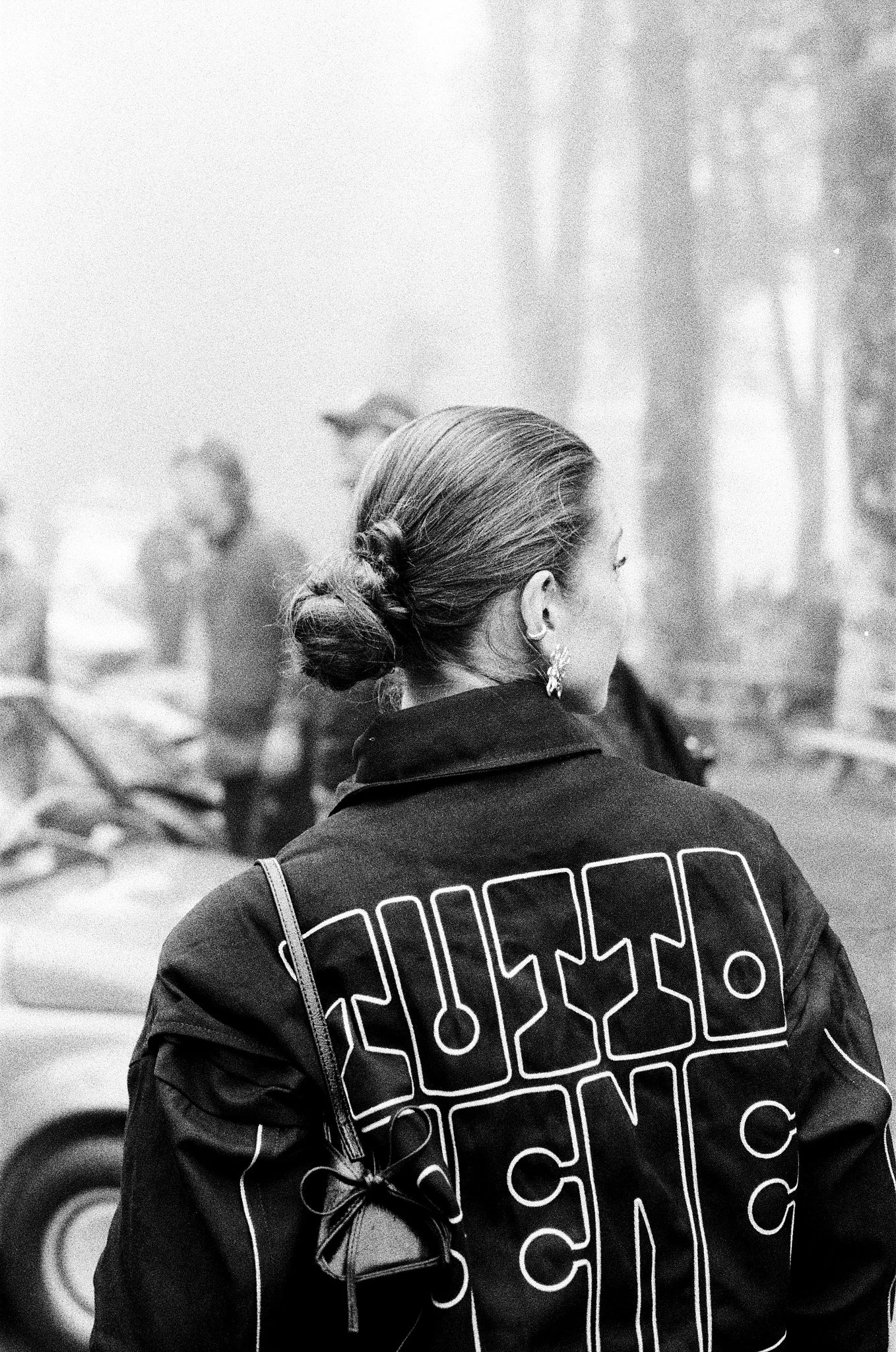 A woman with her hair in a bun, wearing a Tutto Bene jacket, stands in front of a blurred background of other people and cityscape. She has earrings and carries a shoulder bag.