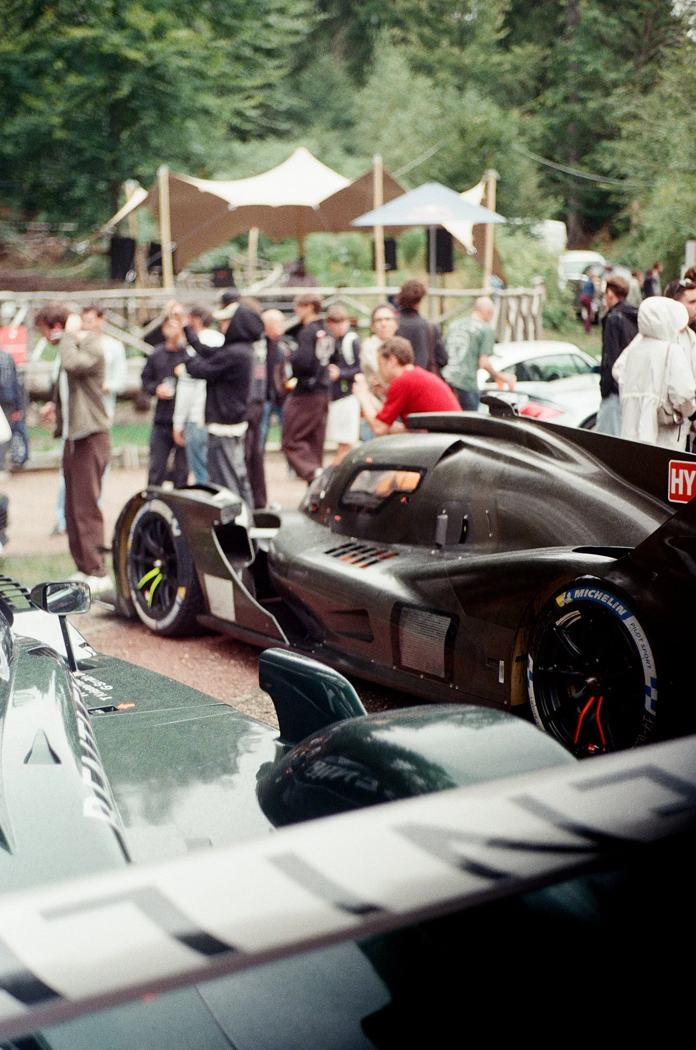 A black race car parked among other vehicles at a car event, with people socializing in the background and trees surrounding the area.