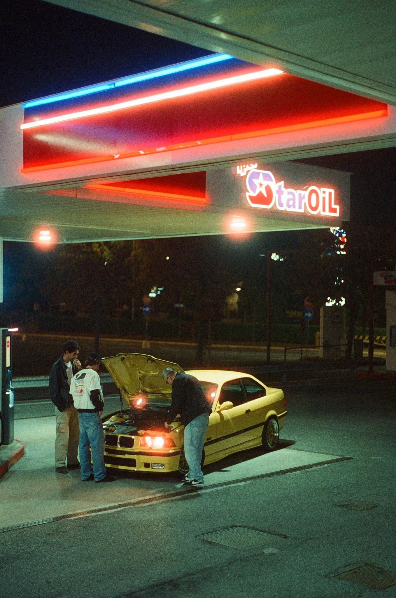 Four men are standing around a yellow car with its hood open at night, outside a gas station with neon signs.