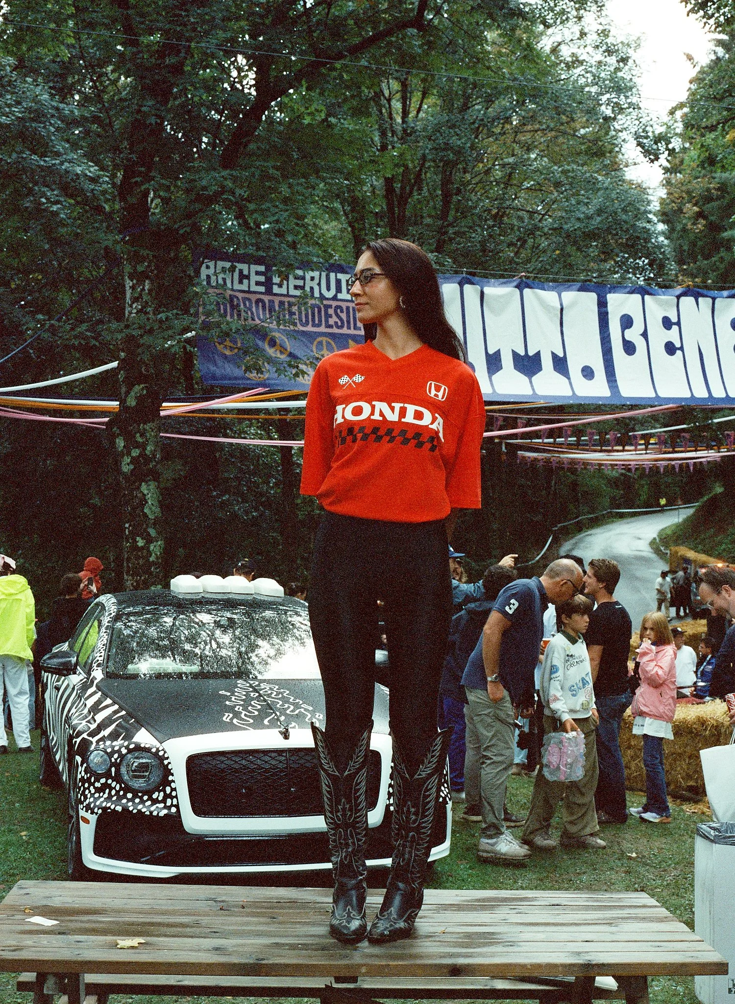A woman with long dark hair, glasses, and earrings stands on a wooden platform in front of a white car with a black and white pattern. She wears a red Honda racing shirt, black pants, and black boots. In the background, a crowd of people and banners 